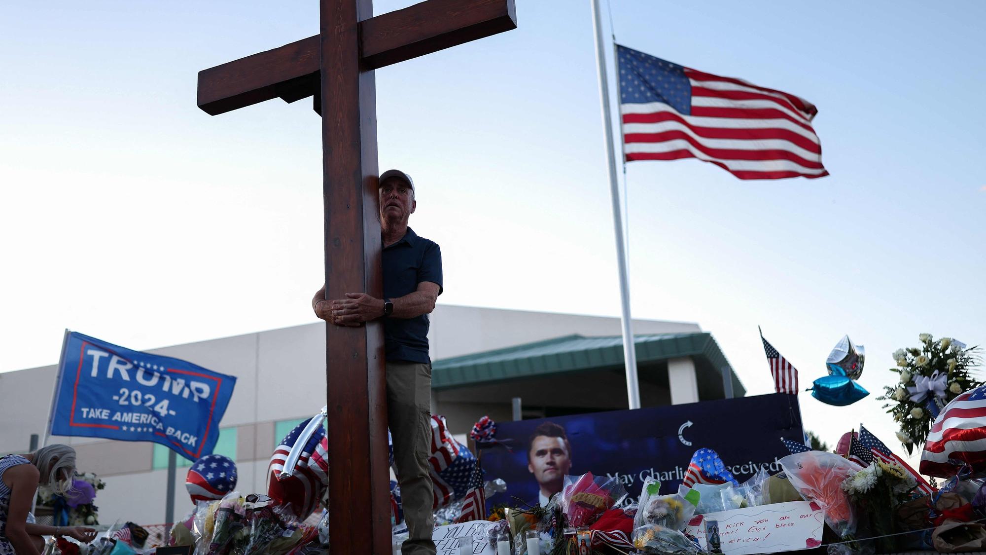TOPSHOT - A US flag flies half-staff as a man carries a large cross near a makeshift memorial for Turning Point USA Founder Charlie Kirk outside of the Turning Point USA headquarters in Phoenix, Arizona, on September 12, 2025. The widow of prominent right-wing activist Charlie Kirk pledged on September 12 to carry on her husband's work, after US authorities announced his alleged assassin had finally been captured. The 31-year-old Kirk was hit by a single bullet while addressing a large crowd at Utah Valley University in the town of Orem on September 10. (Photo by CHARLY TRIBALLEAU / AFP)