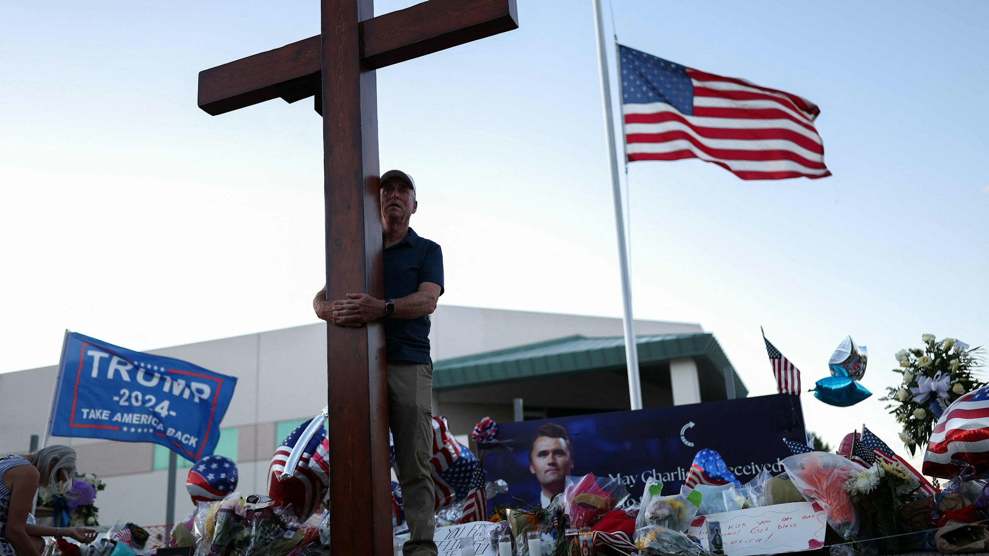 TOPSHOT - A US flag flies half-staff as a man carries a large cross near a makeshift memorial for Turning Point USA Founder Charlie Kirk outside of the Turning Point USA headquarters in Phoenix, Arizona, on September 12, 2025. The widow of prominent right-wing activist Charlie Kirk pledged on September 12 to carry on her husband's work, after US authorities announced his alleged assassin had finally been captured. The 31-year-old Kirk was hit by a single bullet while addressing a large crowd at Utah Valley University in the town of Orem on September 10. (Photo by CHARLY TRIBALLEAU / AFP)