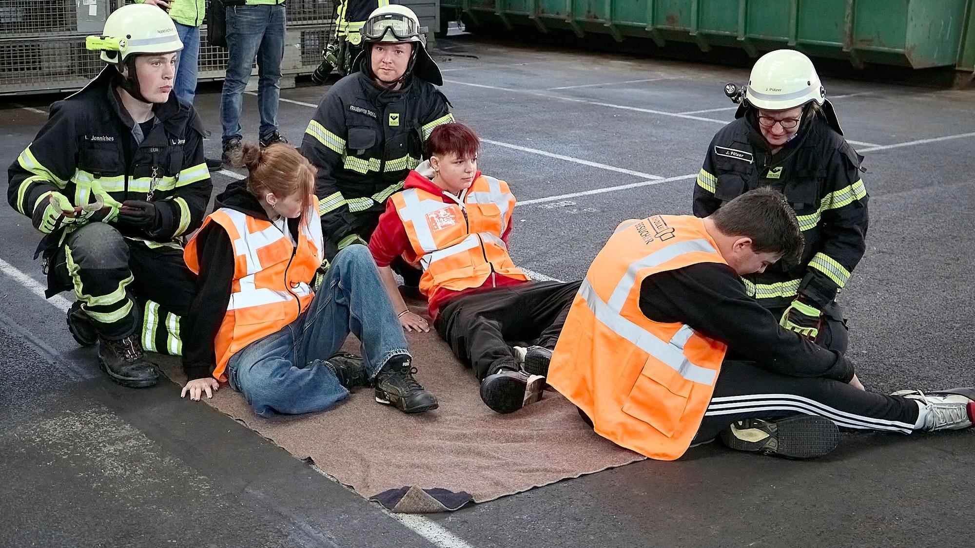 Drei Feuerwehrleute kümmern sich um drei auf einer Rettungsdecke sitzende junge Verletztendarsteller.