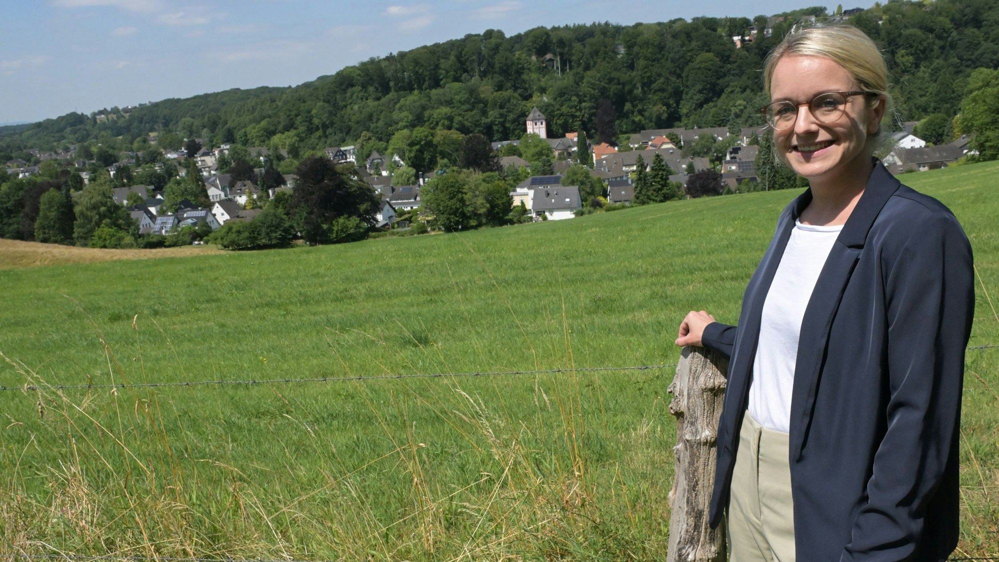 Eine junge Frau mit blonden, glatten Haaren und Brille steht vor einer Wiese mit Blick auf das historische Zentrum von Odenthal.