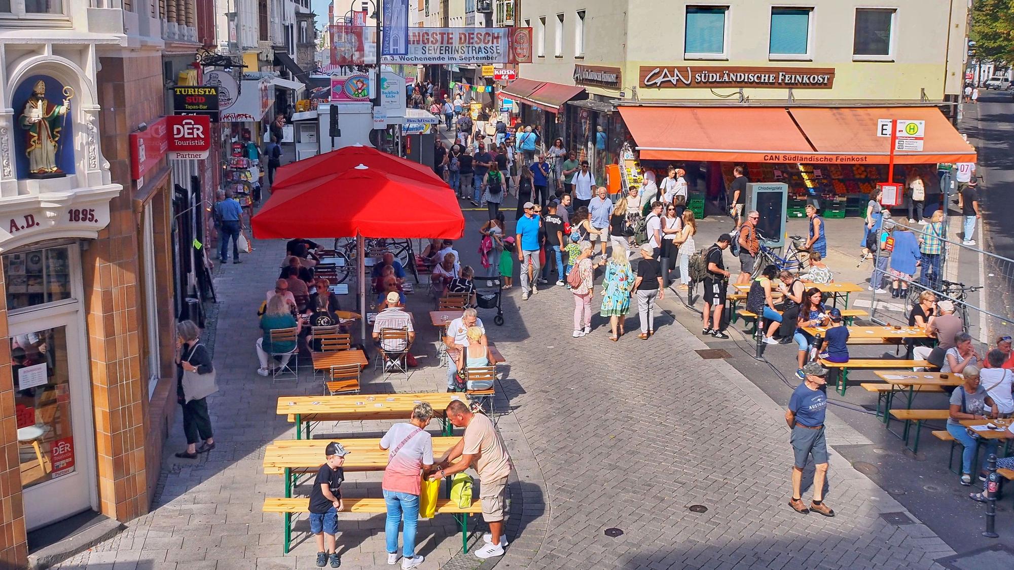 Links und rechst von einer schmalen Straße stehen Bierbänke, Menschen laufen die Straße entlang oder sitzen an den Tischen.