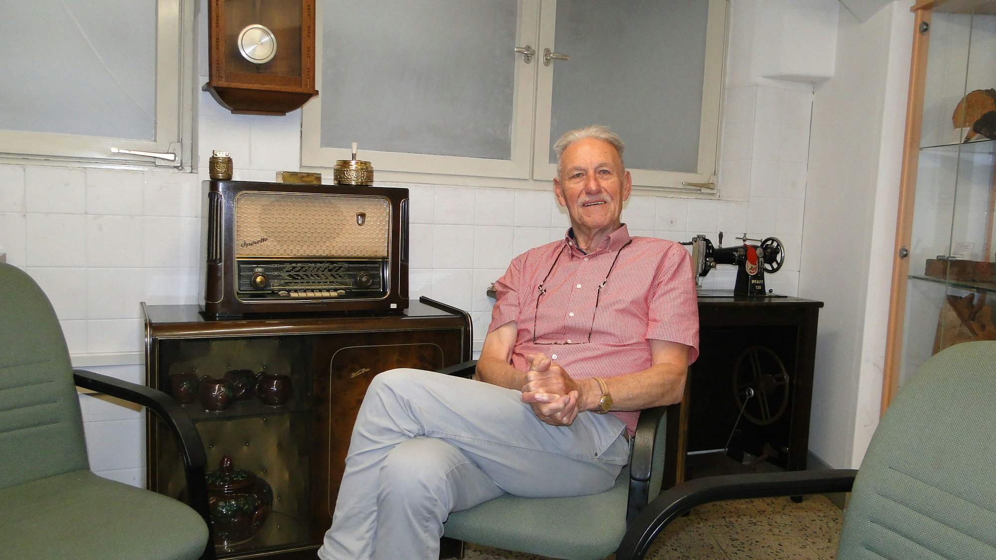 Ein älterer Herr mit grauem Haar und Oberlippenbart sitzt auf einem altmodischen Sessel, neben ihm auf einer Kommode steht ein antikes Radio.