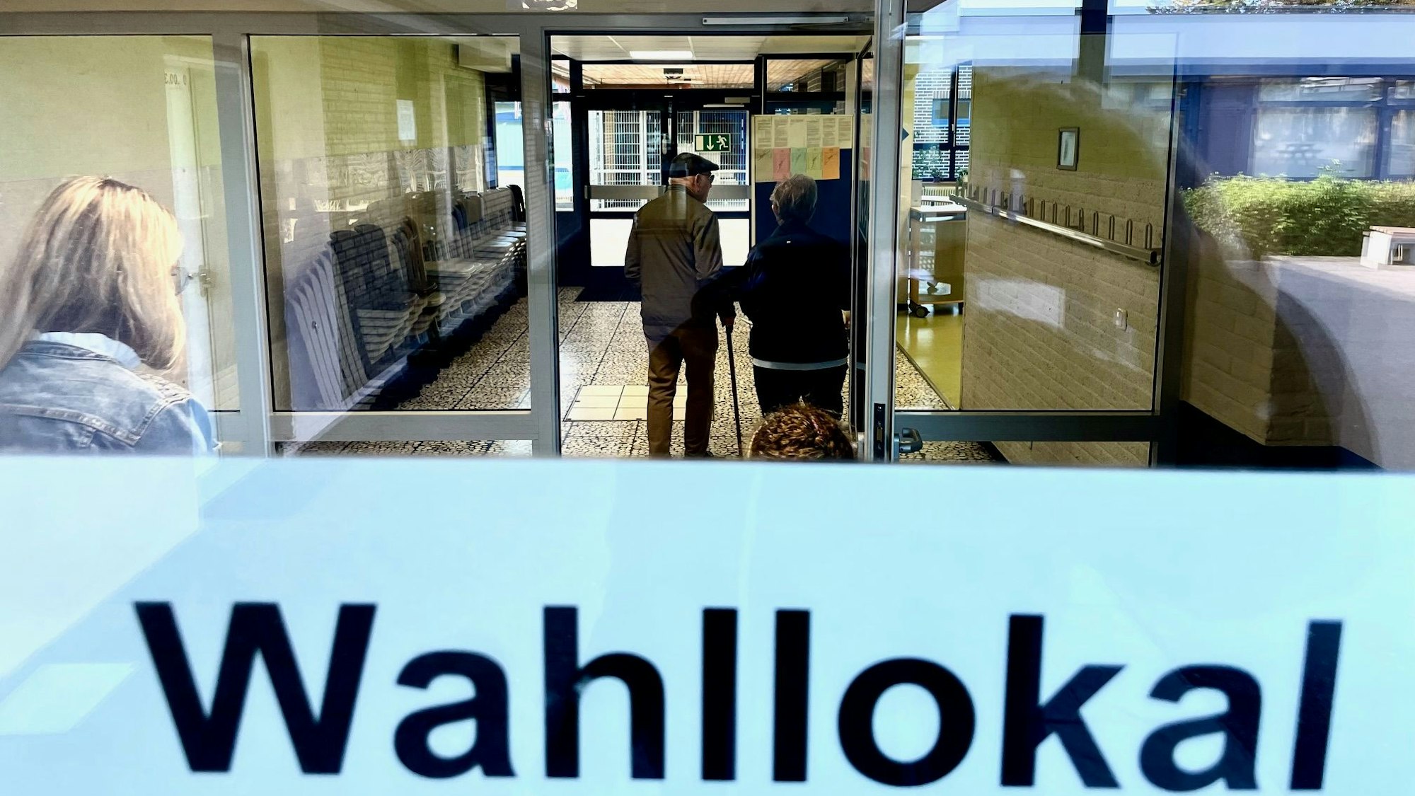 Der Blick geht in einen gefliesten Flur, in dem ein Senior mit einem Stock steht. Auf einer Tür ist der Schriftzug „Wahllokal“.