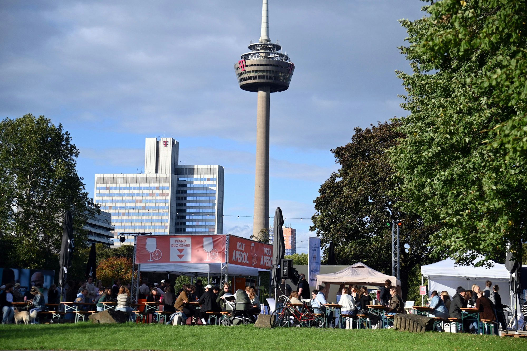 Auf der grünen Wiese zwischen Bäumen stehen Pop-up-Zelte und Bänke, auf denen Menschen sitzen.