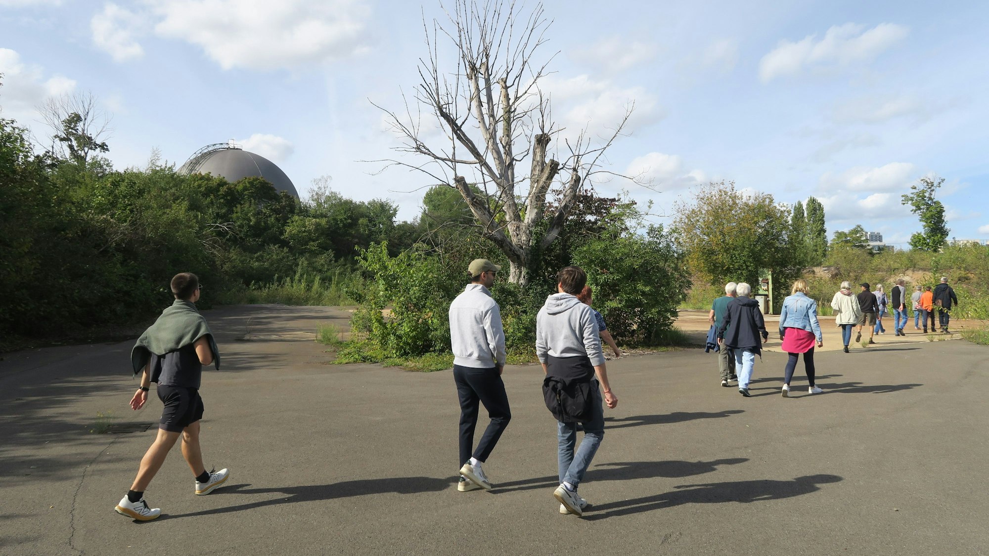 Menschen laufen über ein teils asphaltiertes Gelände, drum herum stehen Bäume und Büsche, eine Gaskugel ist auch zu sehen.