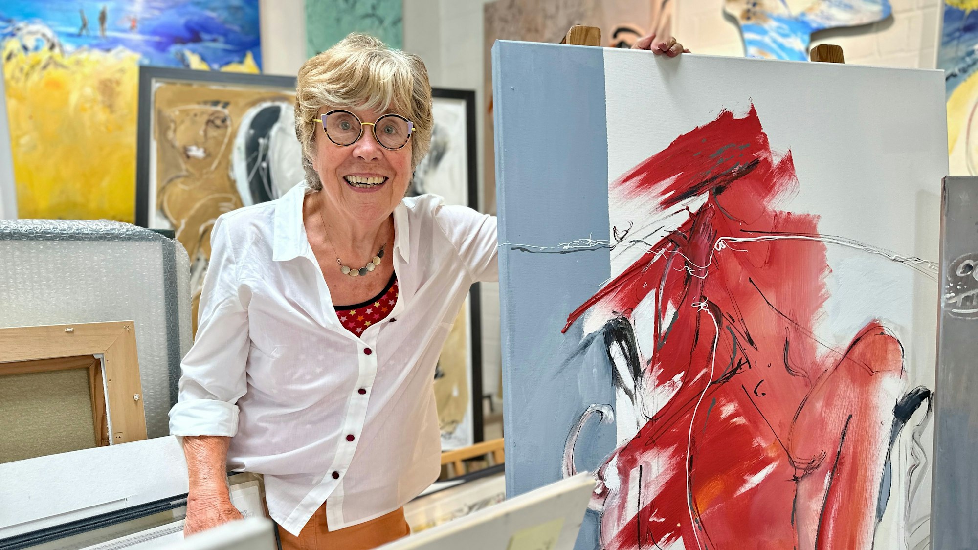 Eine Frau mit Brille und weißer Bluse steht zwischen vielen farbenfrohen Bildern.