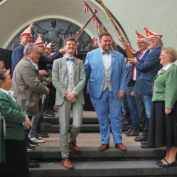 Zwei Männer im Anzug gehen die Stufen einer Kirche herab. Links und rechts stehen Menschen Spalier, teils mit Kappen und Gewehren.