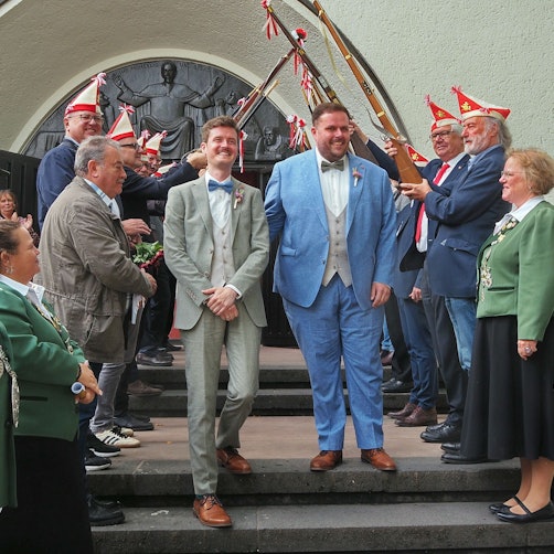 Zwei Männer im Anzug gehen die Stufen einer Kirche herab. Links und rechts stehen Menschen Spalier, teils mit Kappen und Gewehren.