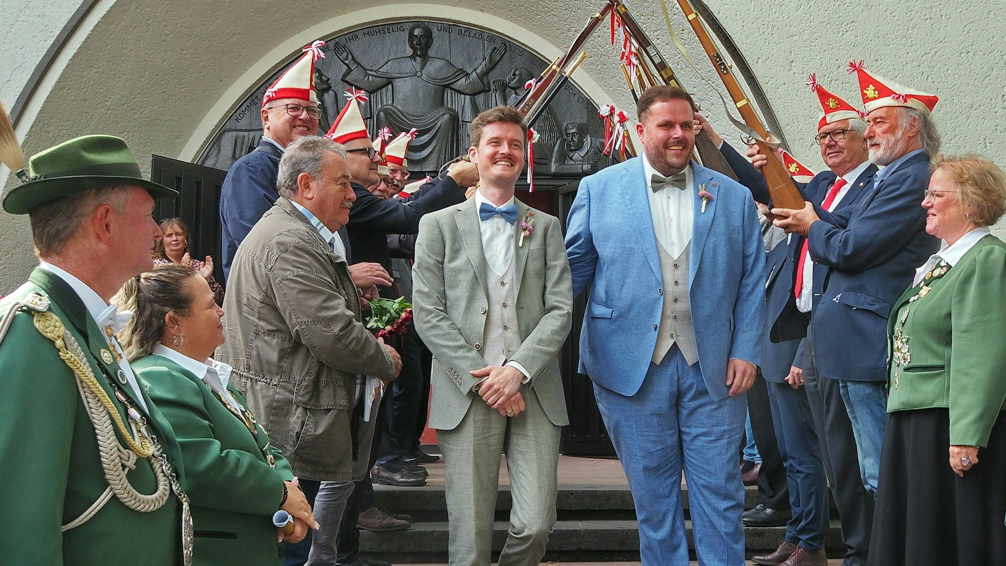Zwei Männer im Anzug gehen die Stufen einer Kirche herab. Links und rechts stehen Menschen Spalier, teils mit Kappen und Gewehren.