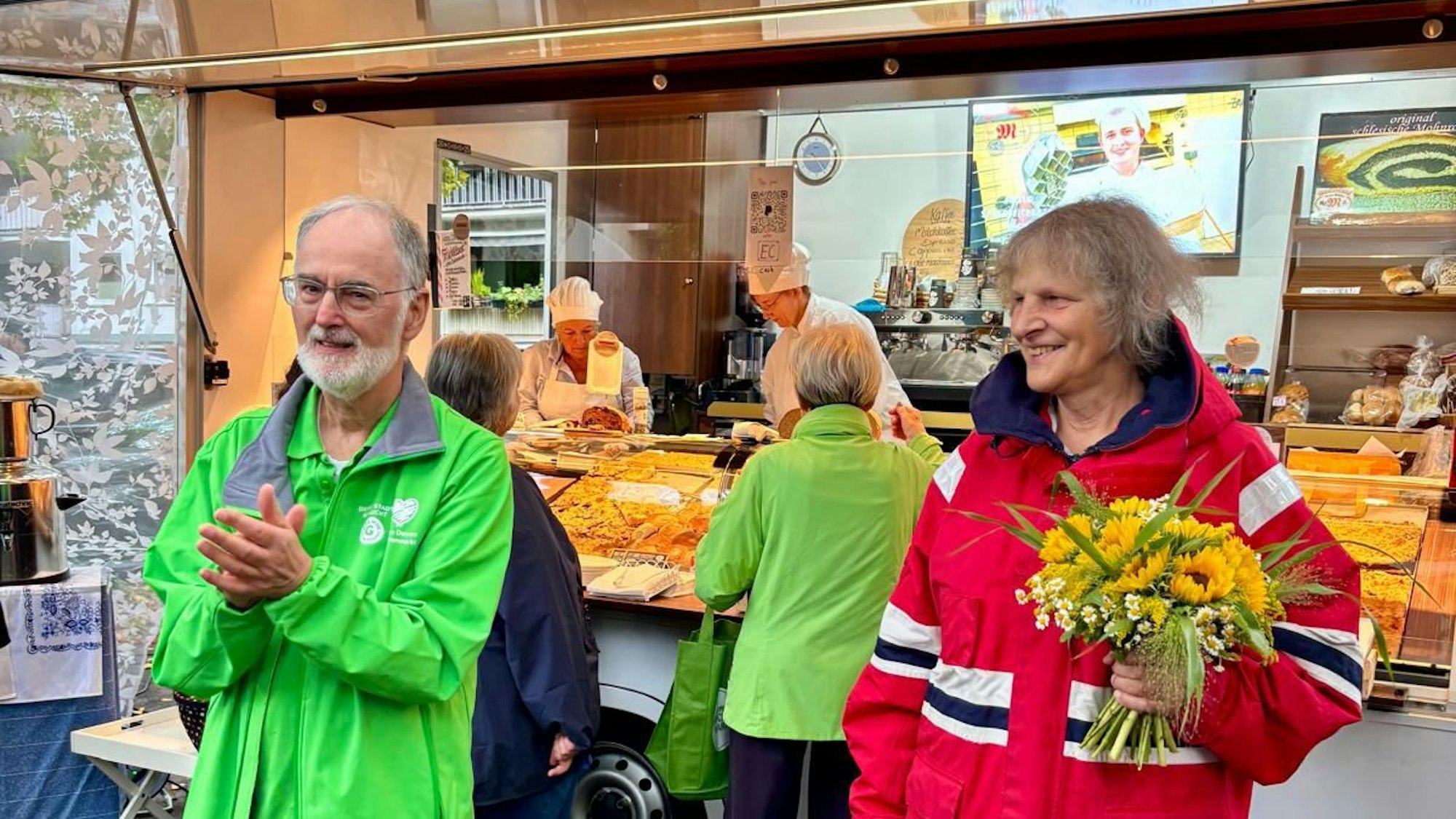 Ein Mann in grüner Jacke und eine Frau in roter Jacke stehen vor einem Bäckereistand auf einem Wochenmarkt.