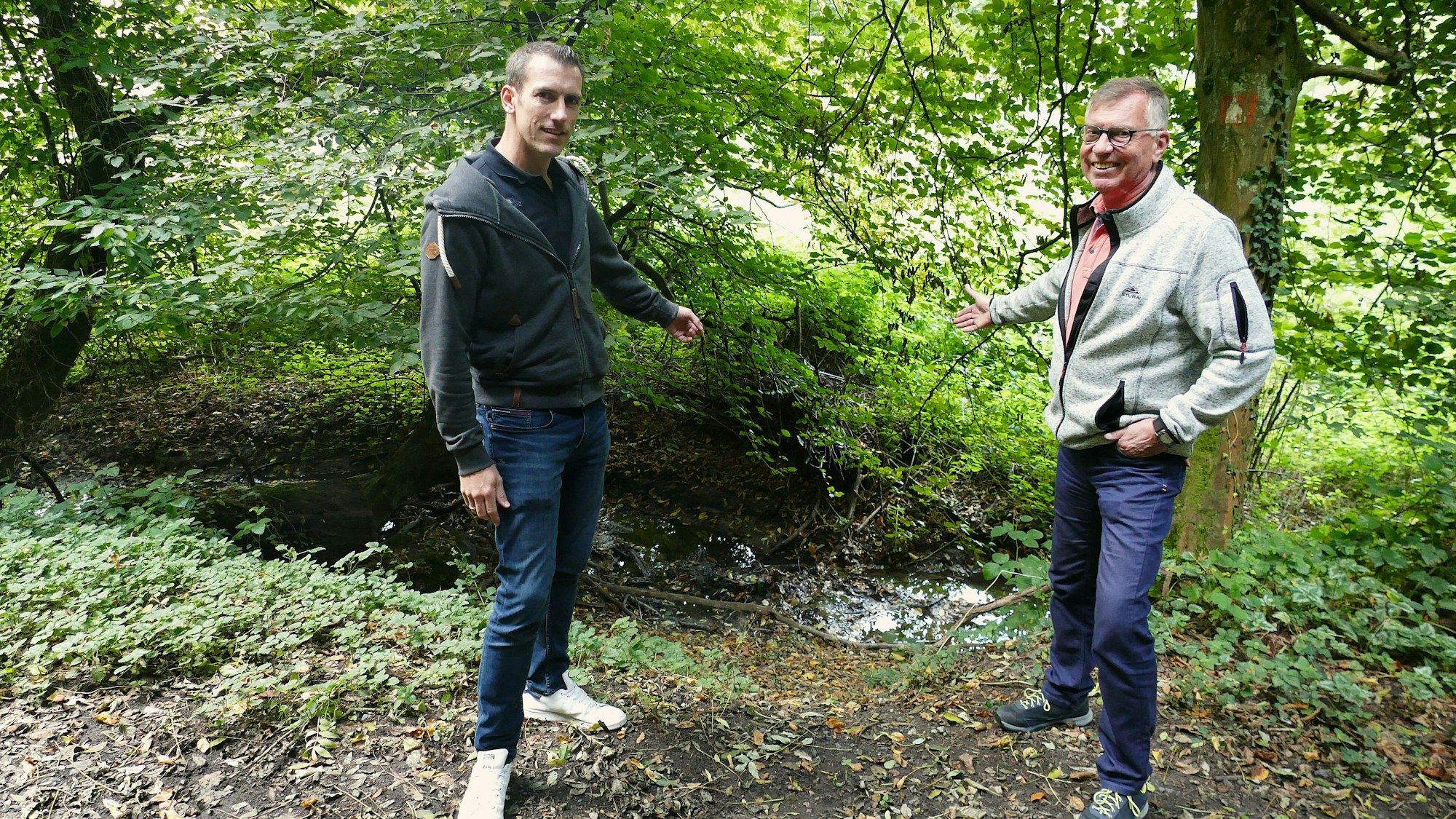 Ein Bach schlängelt sich durch den Wald, umgeben von Uferböschungen und Bäumen. Zwei Männer stehen davor und zeigen auf das Wasser.