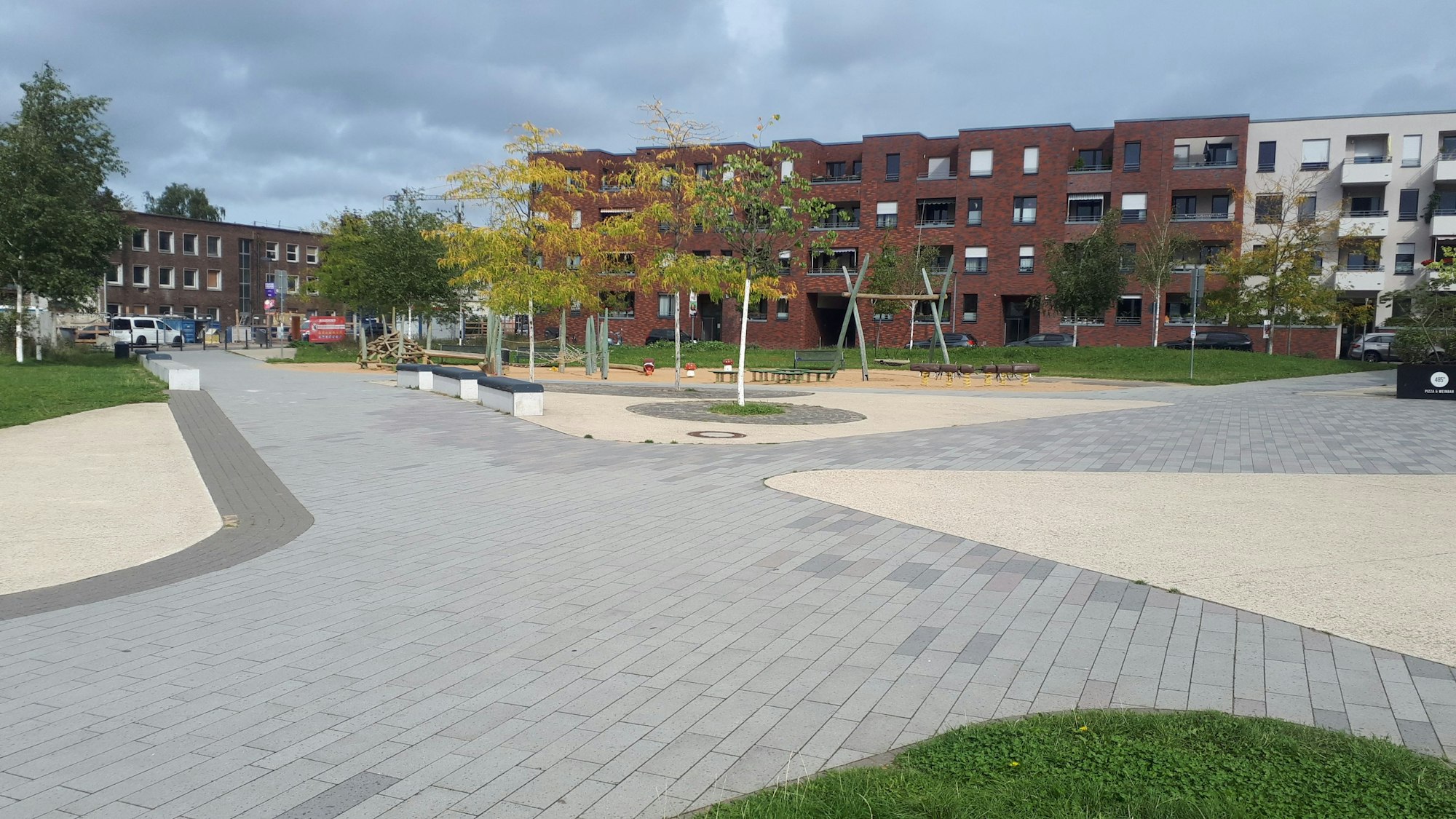 Ein gepflasterter Platz mit ein paar kleinen Bäumen und einem Spielplatz. Im Hintergrund sind Wohnhäuser zu sehen.