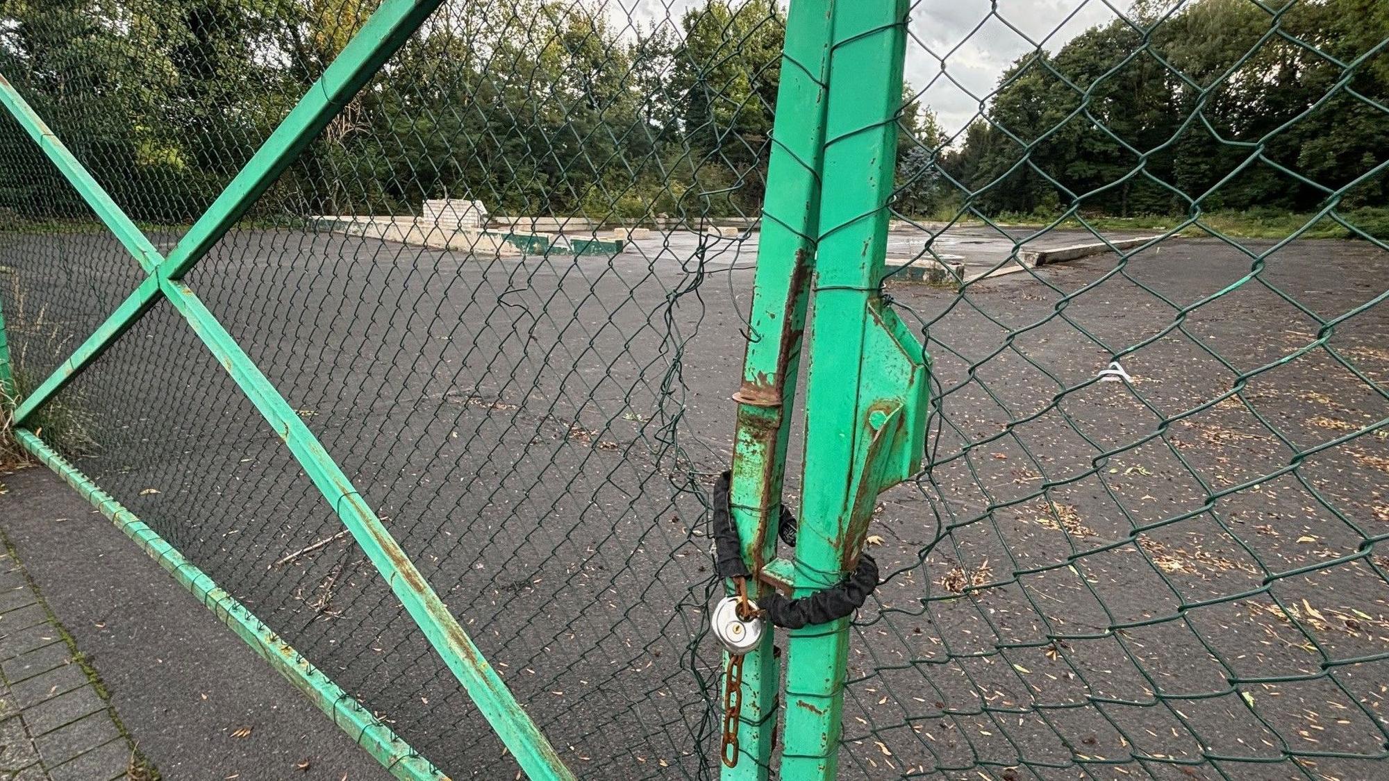 Auf dem Bild ist ein Zaun mit einem Kettenschloss vor einem brachliegenden Grundstück zu sehen.