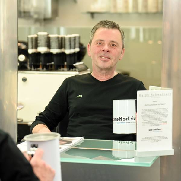 Porträt von Ralph Schwalbach, Großspender von "wir helfen". Er sitzt in seinem Friseursalon vor einem Spiegel und hält eine Spendendose in der Hand.