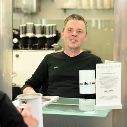 Porträt von Ralph Schwalbach, Großspender von "wir helfen". Er sitzt in seinem Friseursalon vor einem Spiegel und hält eine Spendendose in der Hand.