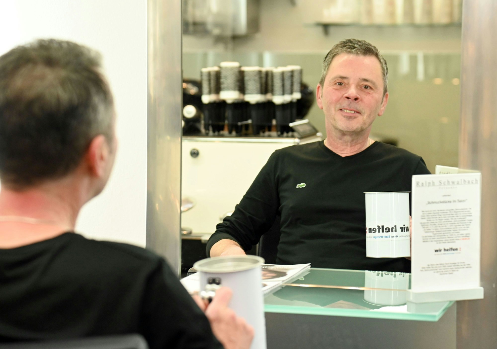 Porträt von Ralph Schwalbach, Großspender von "wir helfen". Er sitzt in seinem Friseursalon vor einem Spiegel und hält eine Spendendose in der Hand.