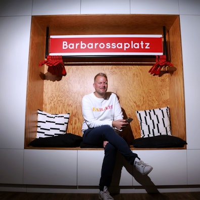 Wilke Stroman sitzt in einer Schrankwandnische - über ihm hängt ein nachgemachtes KVB-Haltestellenschild mit der Aufschrift Barbarossaplatz.