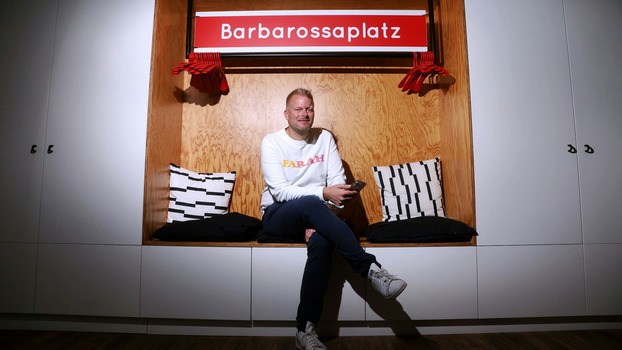 Wilke Stroman sitzt in einer Schrankwandnische - über ihm hängt ein nachgemachtes KVB-Haltestellenschild mit der Aufschrift Barbarossaplatz.