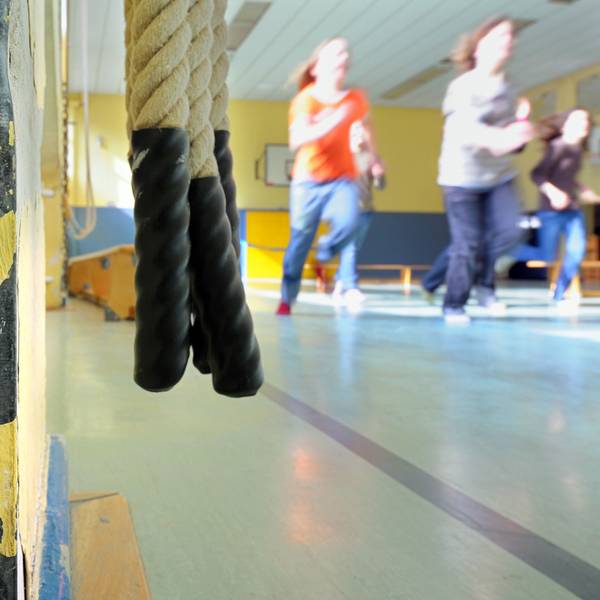 Kinder laufen in einer Turnhalle einer Schule an der Zuleitung eines Heizkörpers vorbei, in dessen Umgebung bereits der Putz abgebröckelt ist.