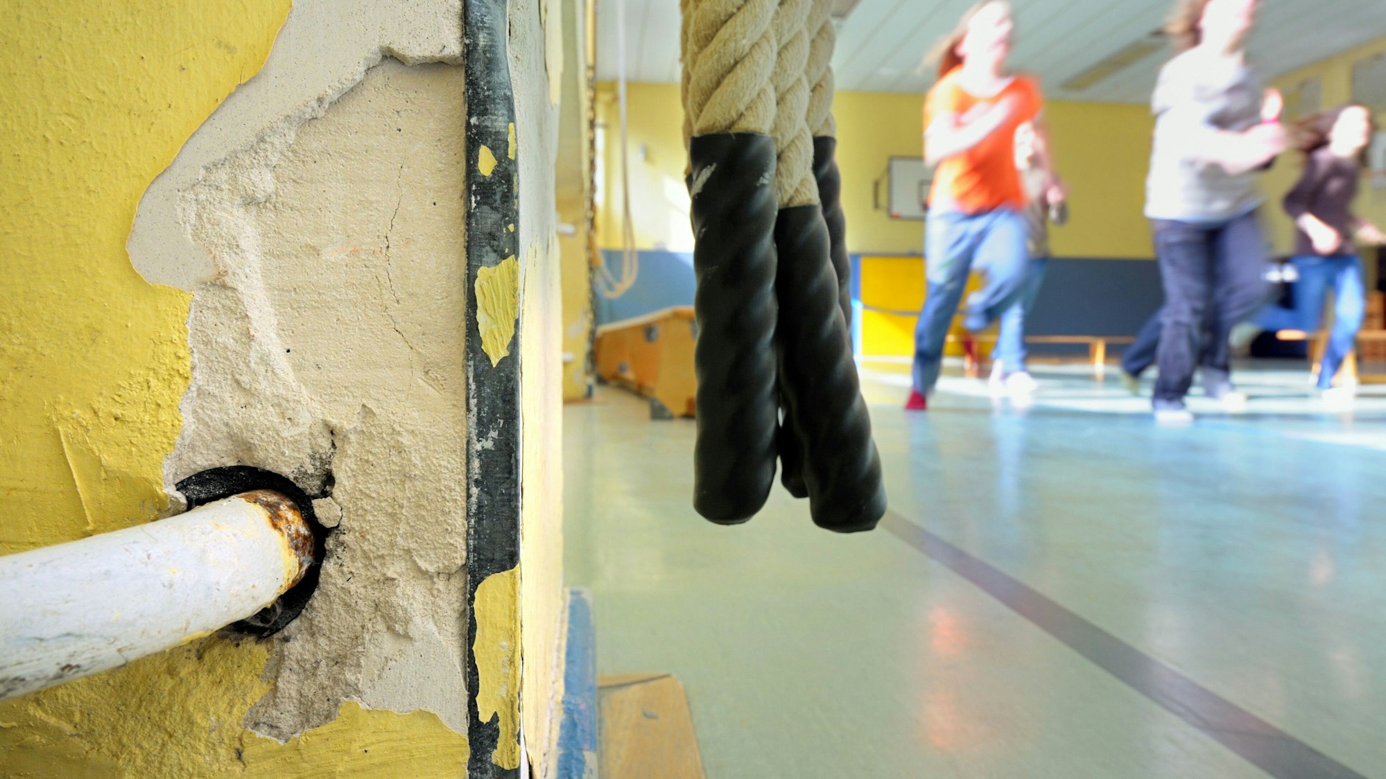 Kinder laufen in einer Turnhalle einer Schule an der Zuleitung eines Heizkörpers vorbei, in dessen Umgebung bereits der Putz abgebröckelt ist.
