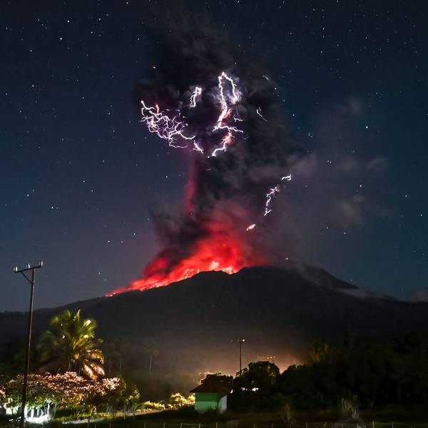Das Bild zeigt den Kegel am Vulkan Lewotobi Laki-Lak. Bei dem Ausbruch steigt eine hohe Aschewolke in den Himmel und glühende Lava rinnt die Hänge hinunter. Durch die längere Belichtungszeit sind die Sterne am Firmament gut zu erkennen.