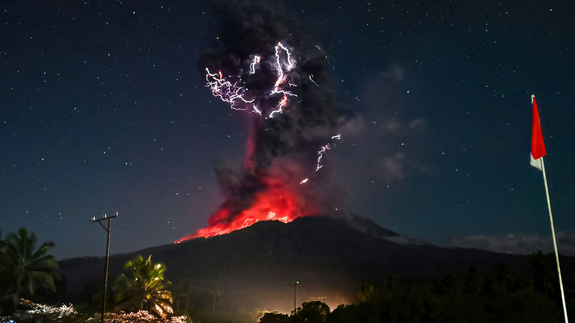 Das Bild zeigt den Kegel am Vulkan Lewotobi Laki-Lak. Bei dem Ausbruch steigt eine hohe Aschewolke in den Himmel und glühende Lava rinnt die Hänge hinunter. Durch die längere Belichtungszeit sind die Sterne am Firmament gut zu erkennen.
