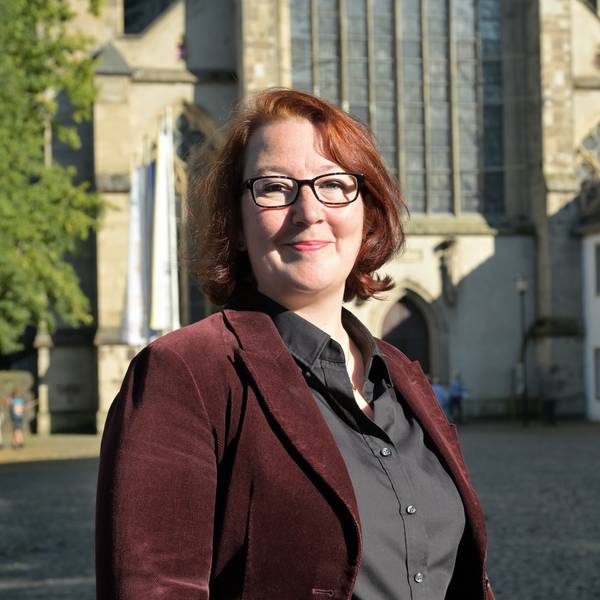 Eine Frau mit rötlichen Haaren und dunkler Brille steht vor dem Kirchenportal des Altenberger Doms.
