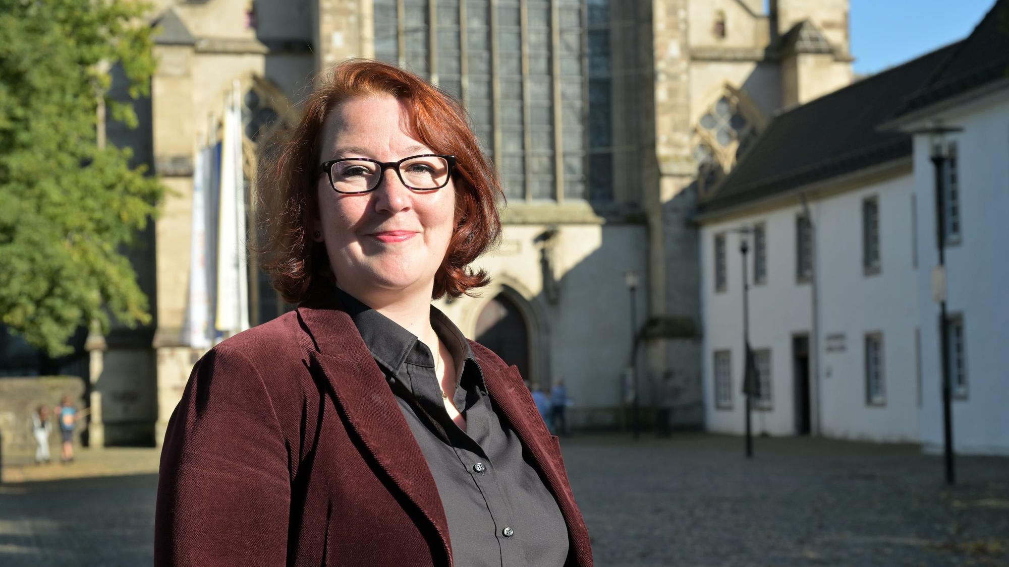 Eine Frau mit rötlichen Haaren und dunkler Brille steht vor dem Kirchenportal des Altenberger Doms.