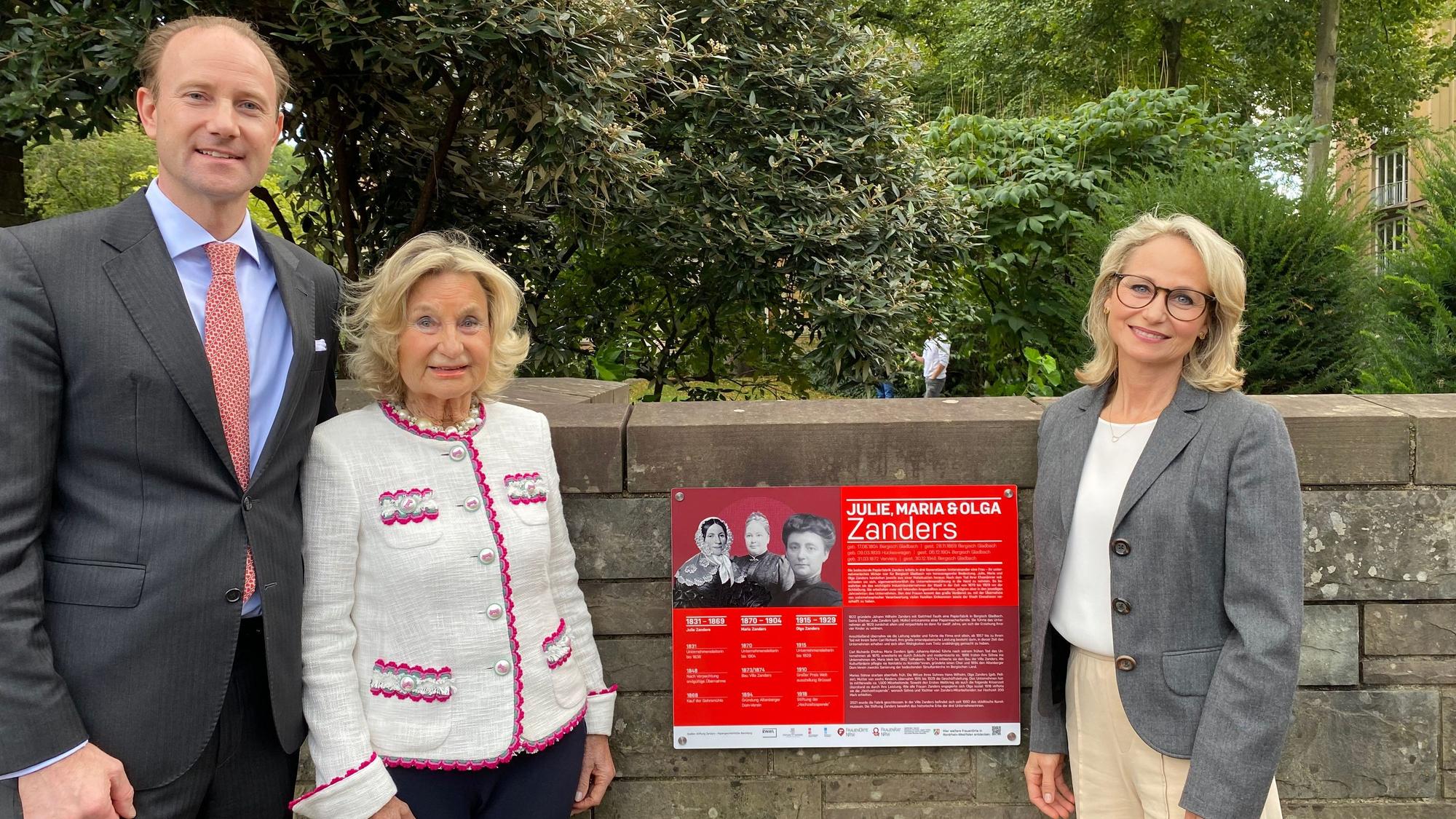 Ein Mann und zwei blonde Frauen vor einer roten Gedenktafel für drei Frauen, die in der Vergangenheit die ehemalige Papierfabrik Zanders geleitet haben.
