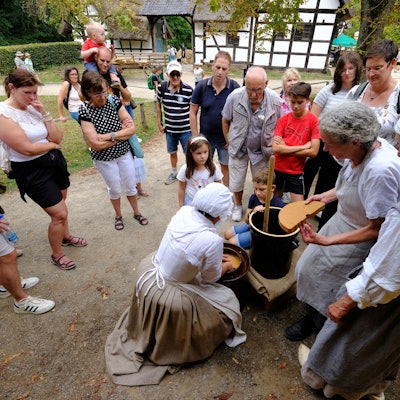 Besucher des Freilichtmuseums schauen den traditionell mit Kopftuch und Schürzen gekleideten Hauswirtschafterinnen bei ihrer Arbeit zu.