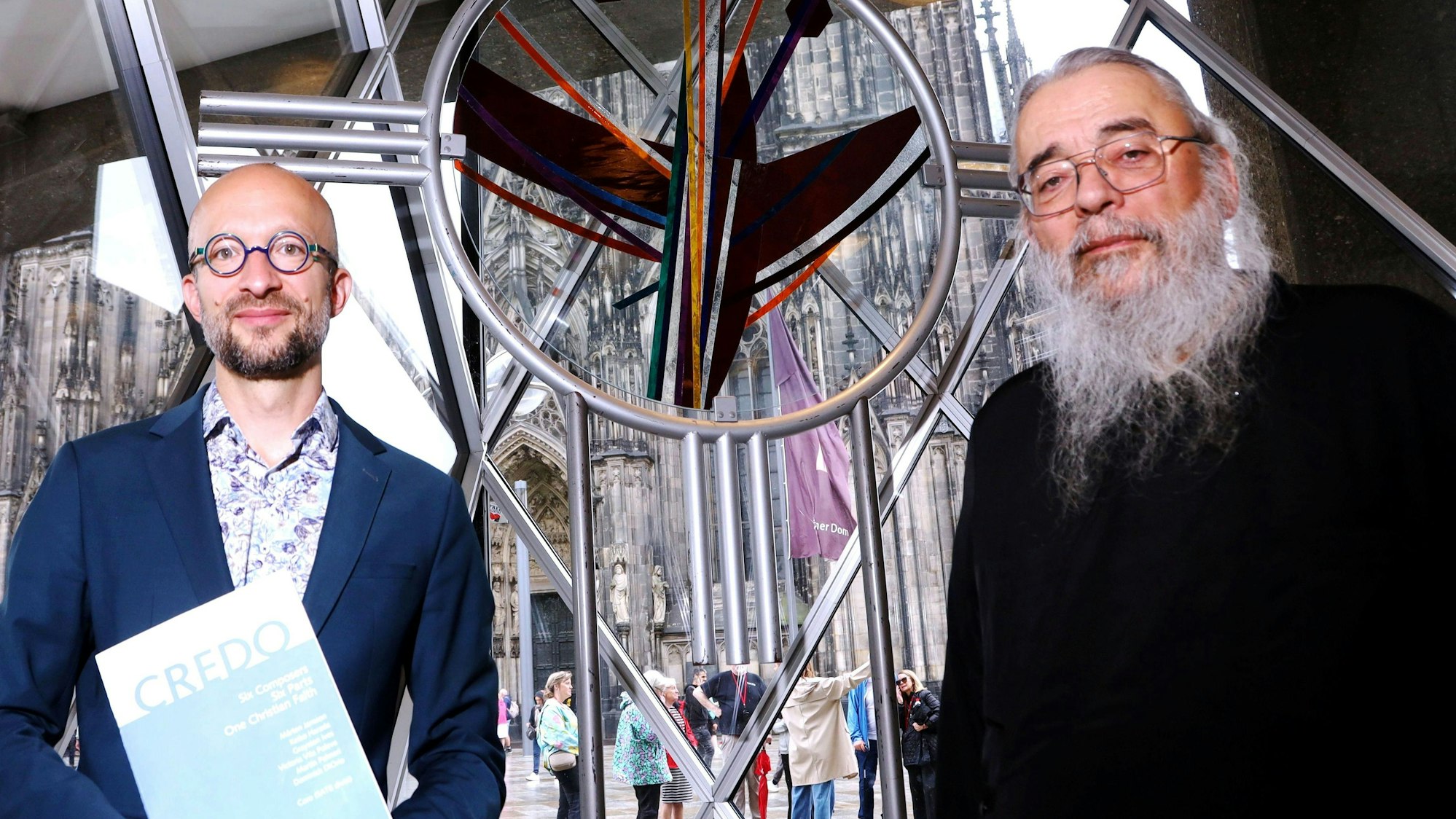 Das Bild zeigt den Kreiskantor Samuel Dobernecker (links) und den griechisch-orthodoxen Erzpriester Radu Constantin Miron zwischen dem für das Domjubiläum 1998 geschaffene Kölner Ökumene- und Versöhnungskreuz des Künstlers Rafael Seitz im Domforum. Foto: Arton Krasniqi