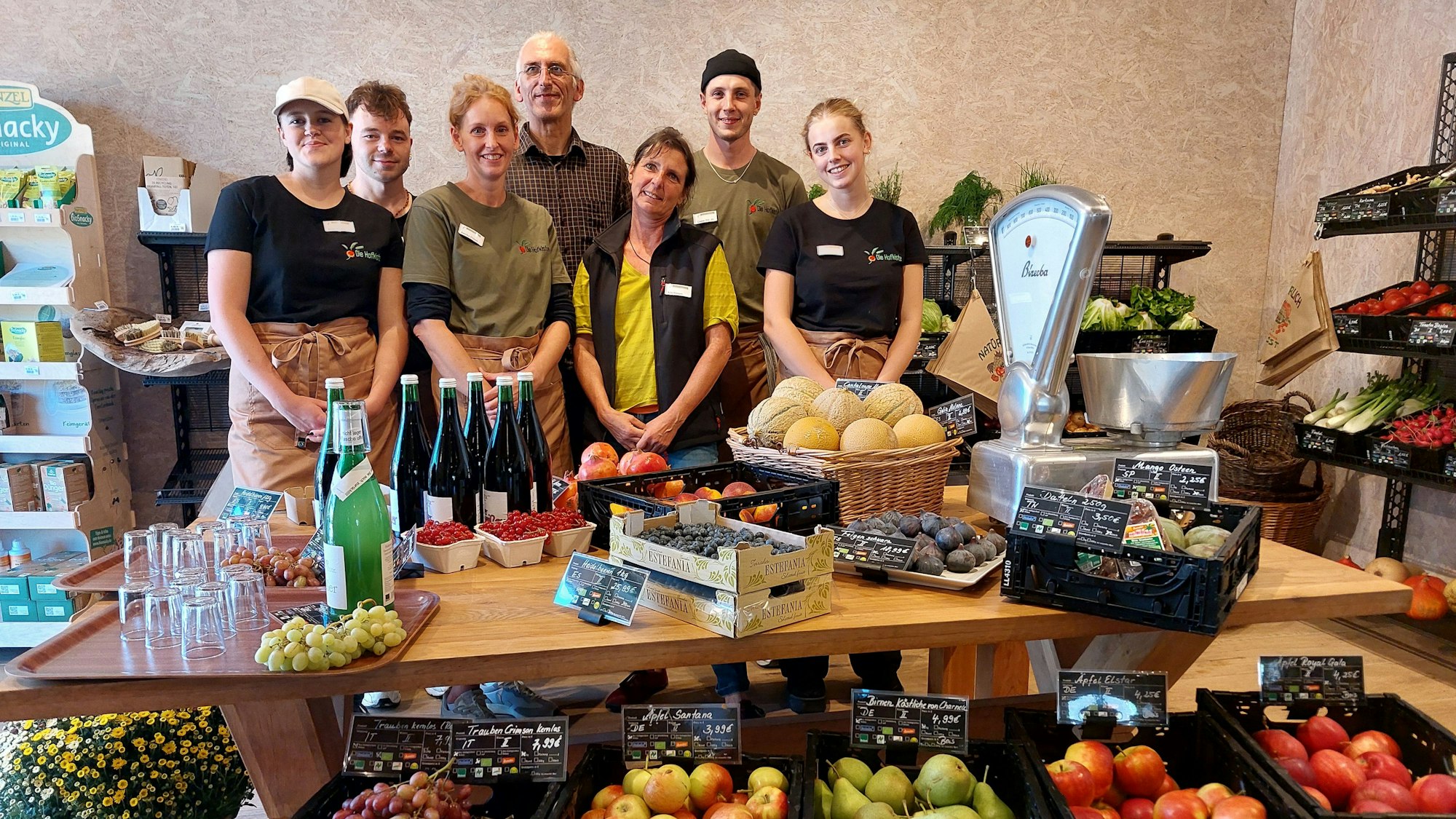 Männer und Frauen stehen hinter einem Tisch mit Obst, Gemüse und einer mechanischen Waage.