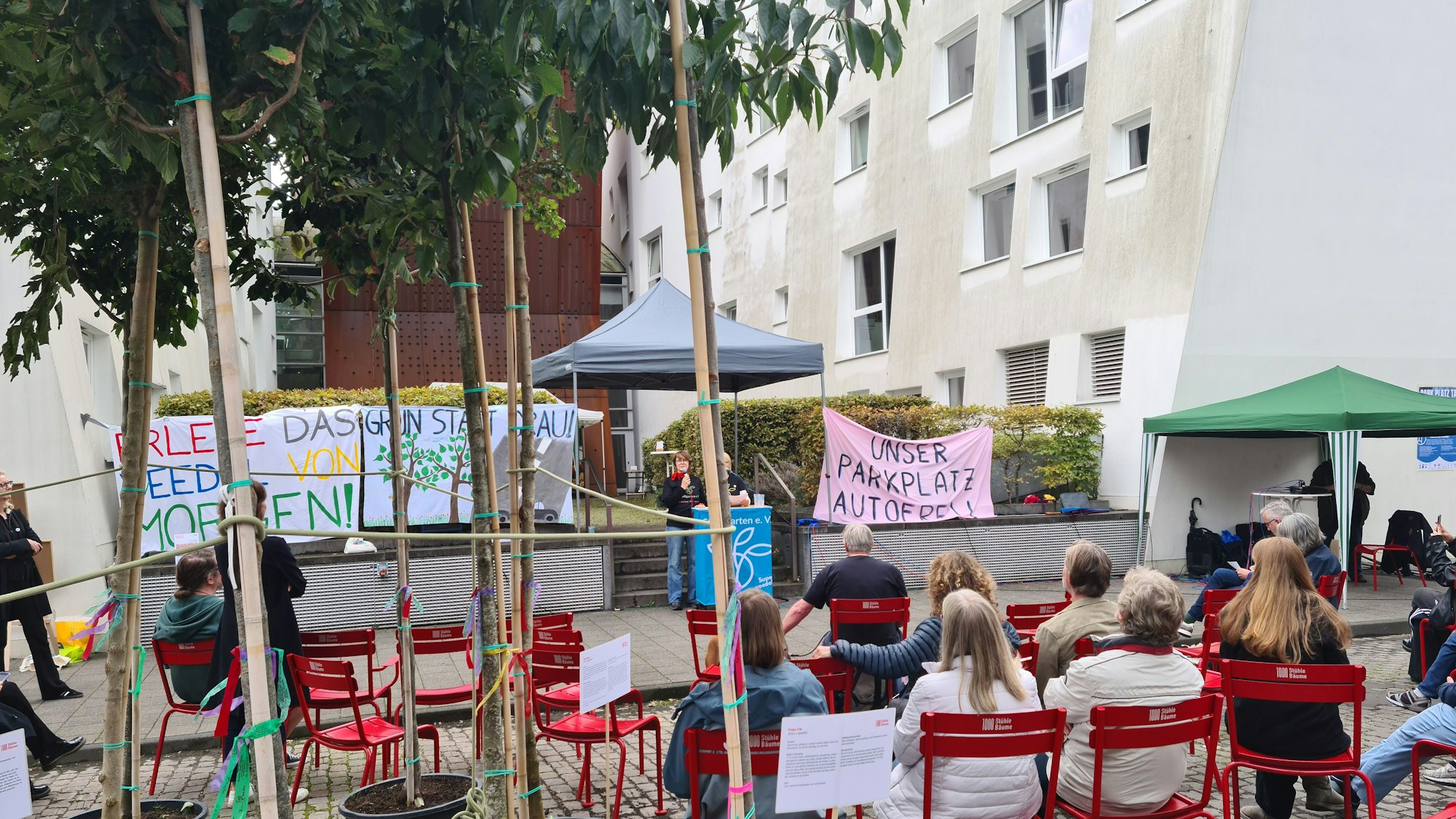 Auf einem Platz sitzen Zuhörer auf roten Stühlen. An einem Rednerpult stehen Menschen, auch Bäume in Plastikpflanzkübeln stehen auf dem Platz.