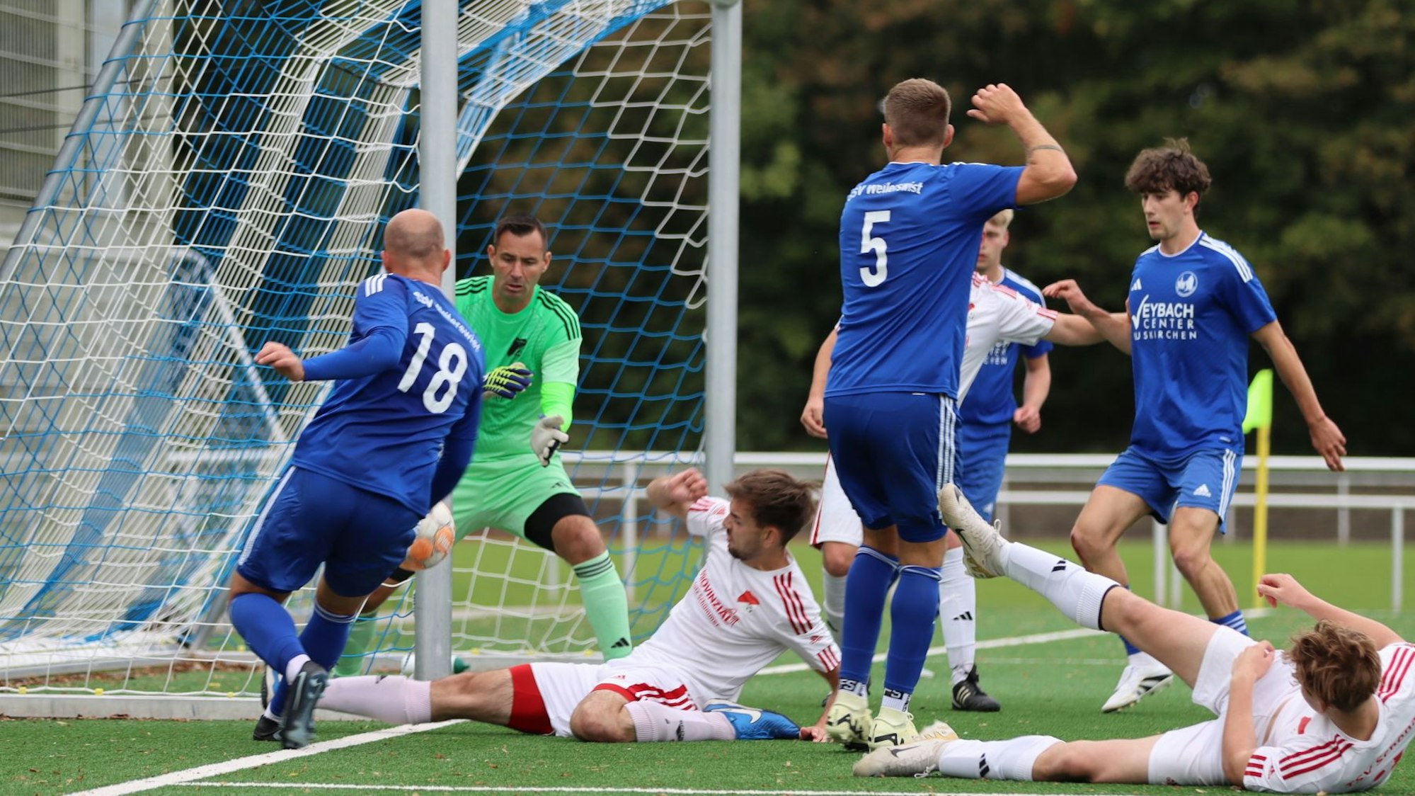 Das Bild zeigt zwei Spieler des TSV Schönau, die auf dem Boden liegen. Ein Spieler des SSV Weilerswist versucht, den Ball nach innen zu spielen.