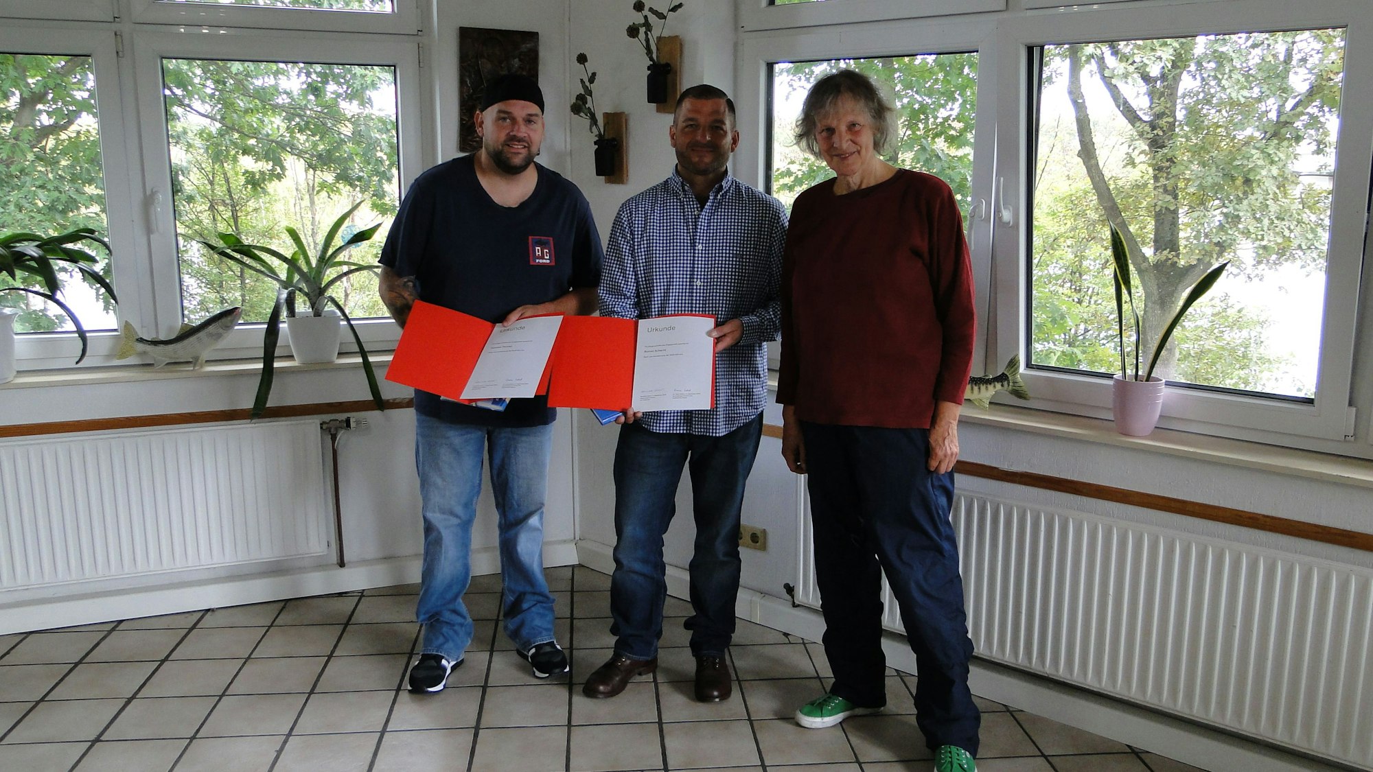 Zwei Männer und eine Frau stehen in einem Raum mit gefliesten Boden und vielen Fenstern. Die Männer halten orangefarbene Kladden mit einer Urkunde in der Hand.