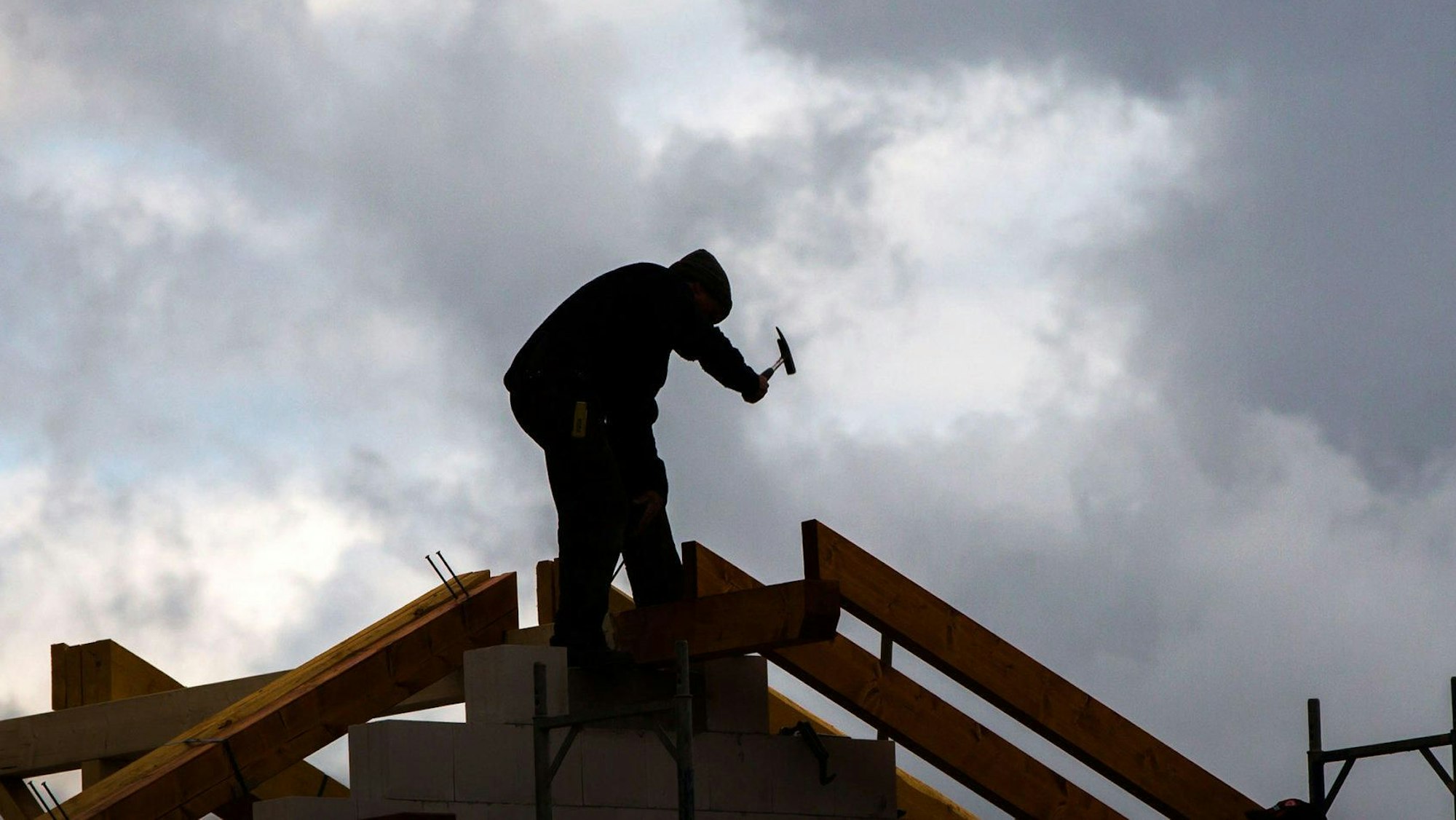 Ein Zimmermann hämmert am Dachstuhl eines im Bau befindlichen Einfamilienhauses, während im Hintergrund graue Regenwolken am Himmel zu sehen sind.