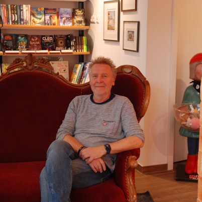 Josef Mütter sitzt in seiner Buchhandlung in Bad Münstereifel auf einem roten Sofa. Rechts ist die Säule aus Salzsteinen zu sehen.