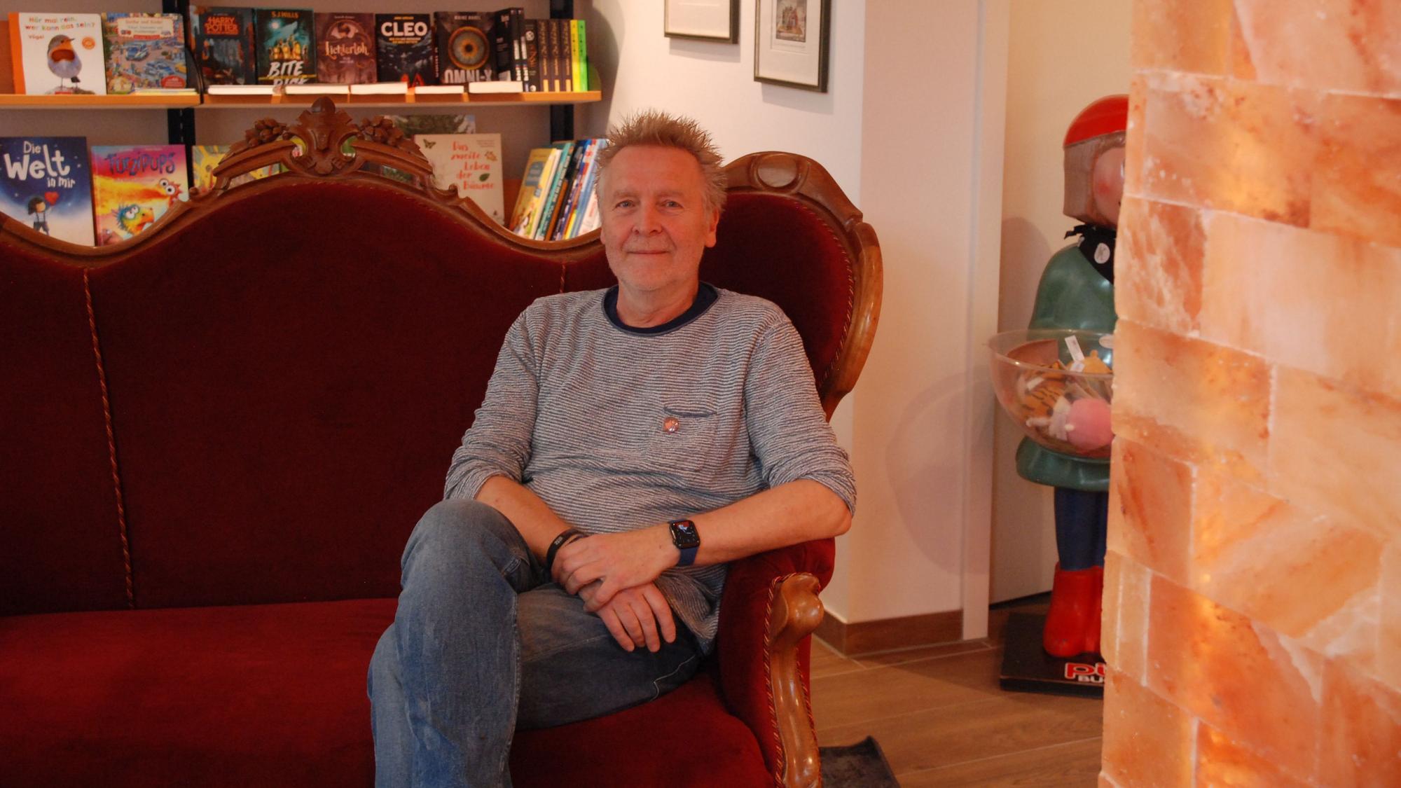 Josef Mütter sitzt in seiner Buchhandlung in Bad Münstereifel auf einem roten Sofa. Rechts ist die Säule aus Salzsteinen zu sehen.