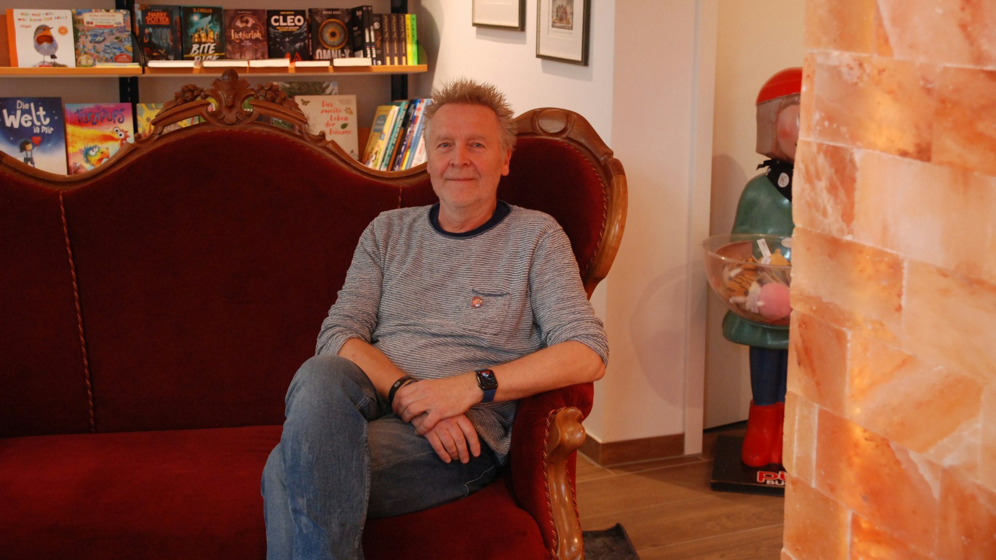 Josef Mütter sitzt in seiner Buchhandlung in Bad Münstereifel auf einem roten Sofa. Rechts ist die Säule aus Salzsteinen zu sehen.
