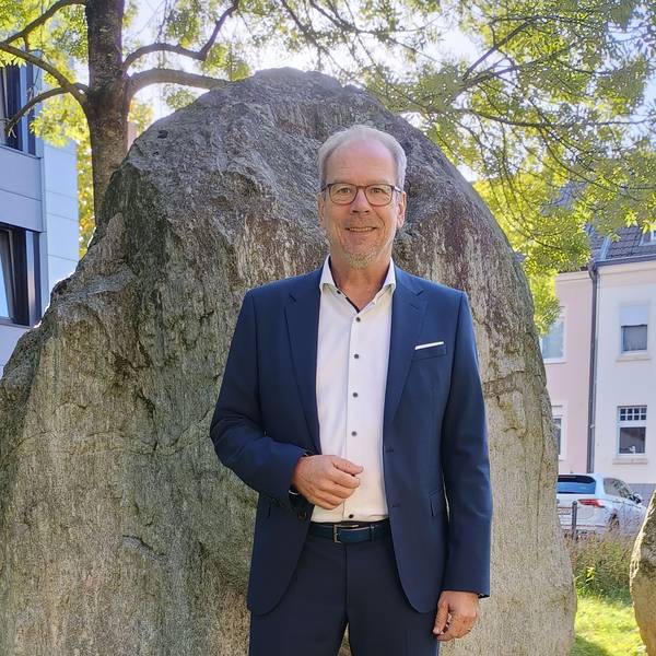 Volker Mießeler steht vor einem großen Stein vor dem Rathaus. Er trägt einen blauen Anzug, ein weißes Hemd und eine Brille.