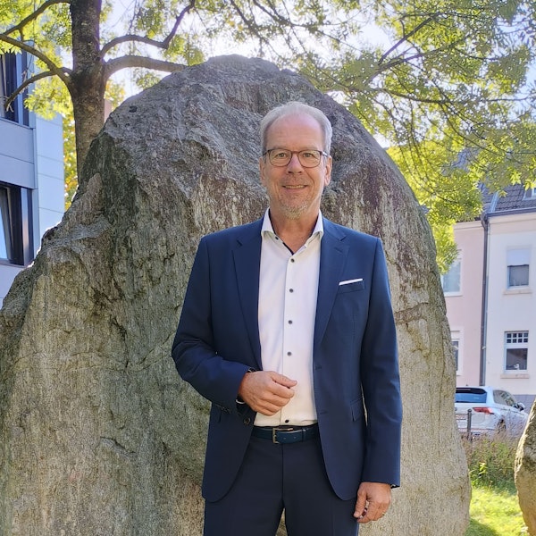 Volker Mießeler steht vor einem großen Stein vor dem Rathaus. Er trägt einen blauen Anzug, ein weißes Hemd und eine Brille.