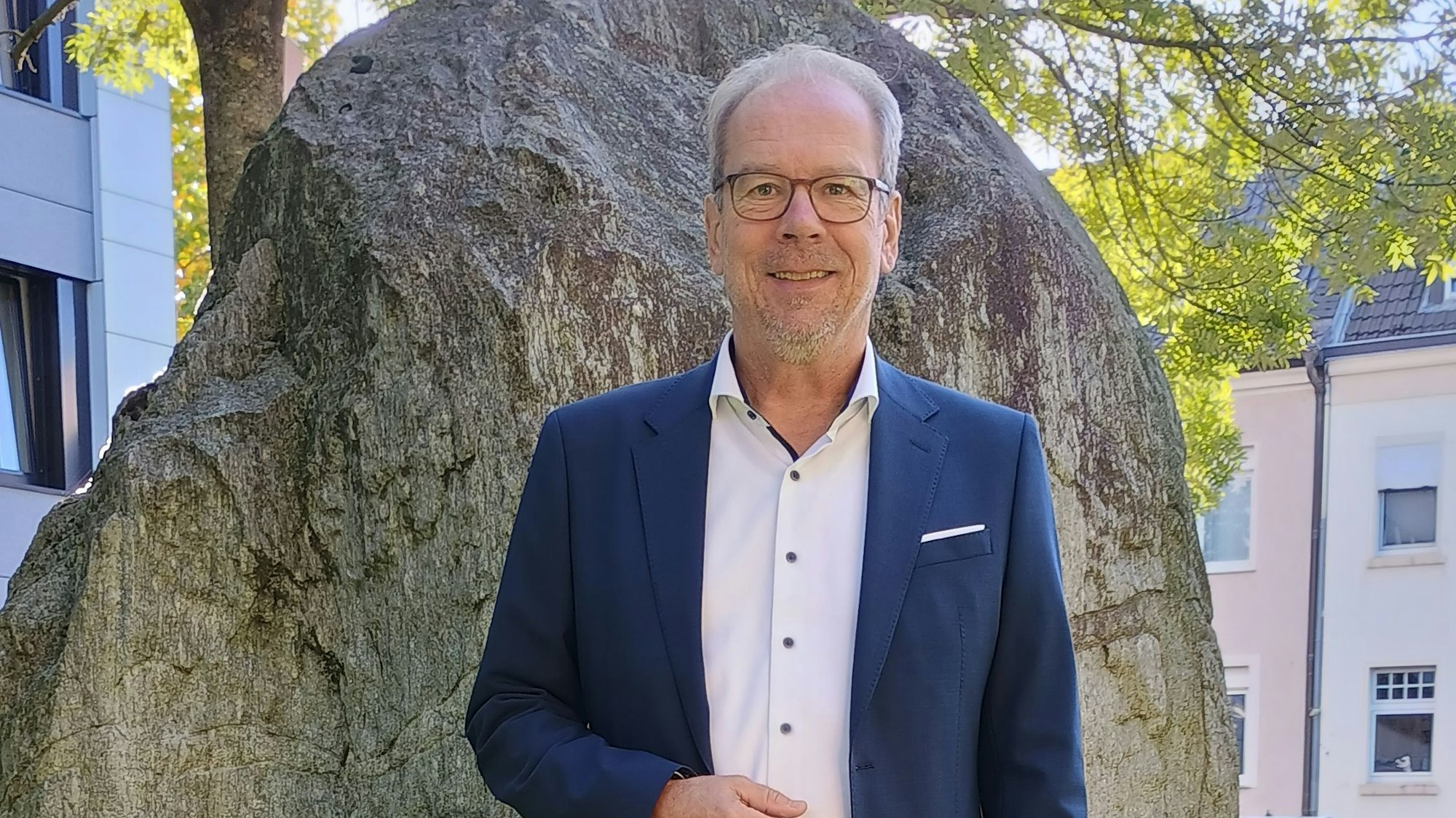 Volker Mießeler steht vor einem großen Stein vor dem Rathaus. Er trägt einen blauen Anzug, ein weißes Hemd und eine Brille.