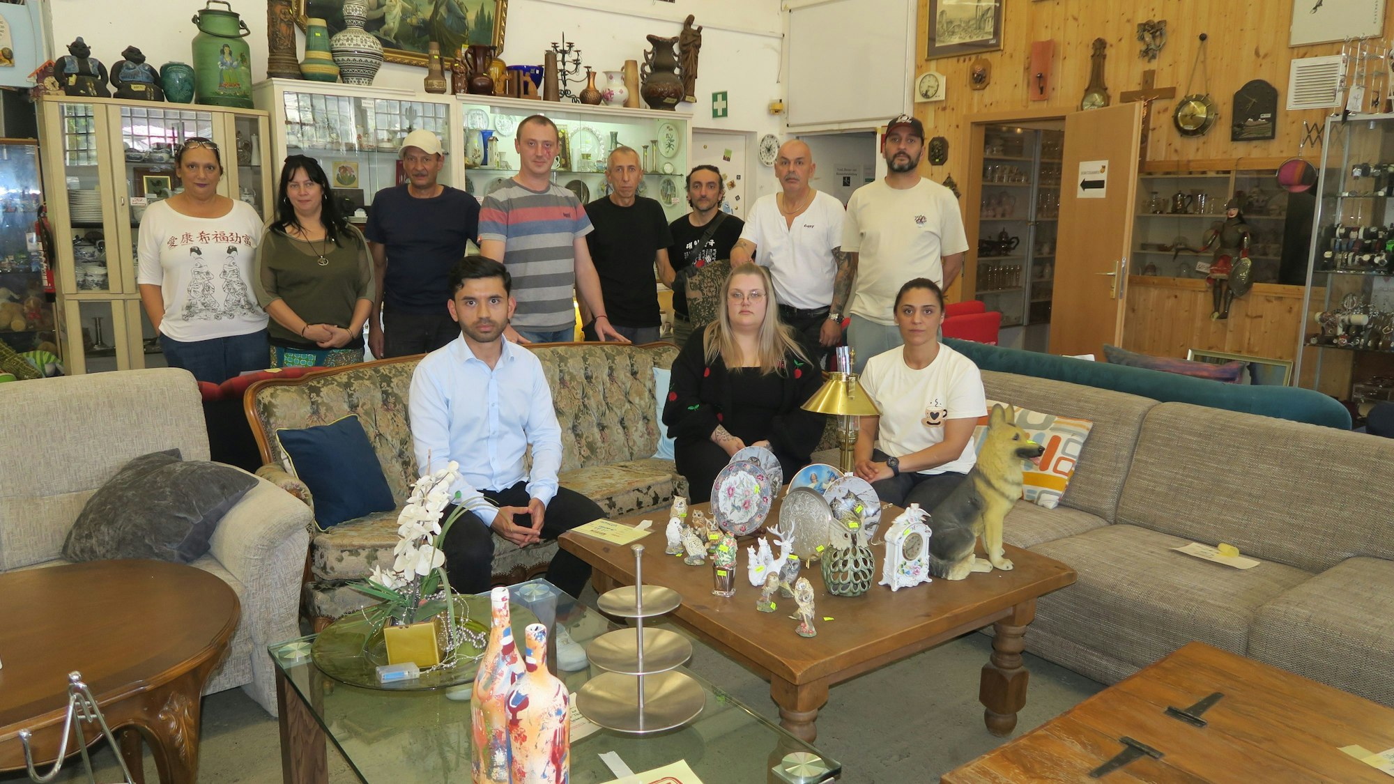 Eine Gruppe Menschen steht und sitzt auf Stühlen in einem großen Raum mit Möbeln und Nippes.