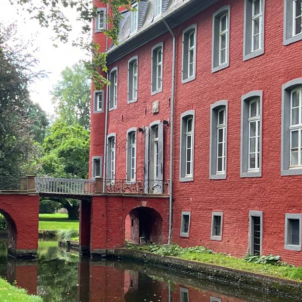 Zu sehen ist ein rotes Schloss mit einer Brücke über einen Schlossgraben.