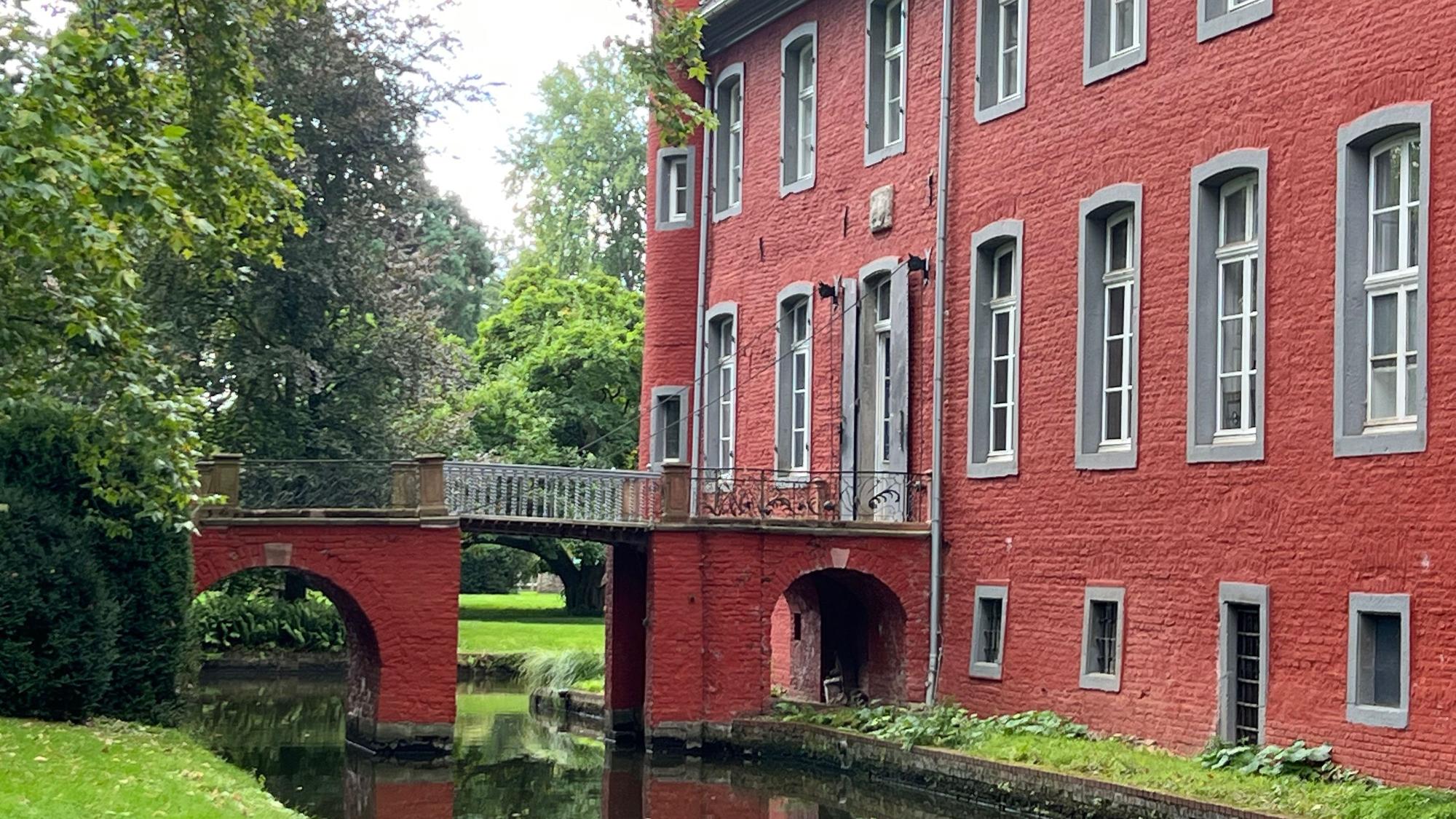 Zu sehen ist ein rotes Schloss mit einer Brücke über einen Schlossgraben.