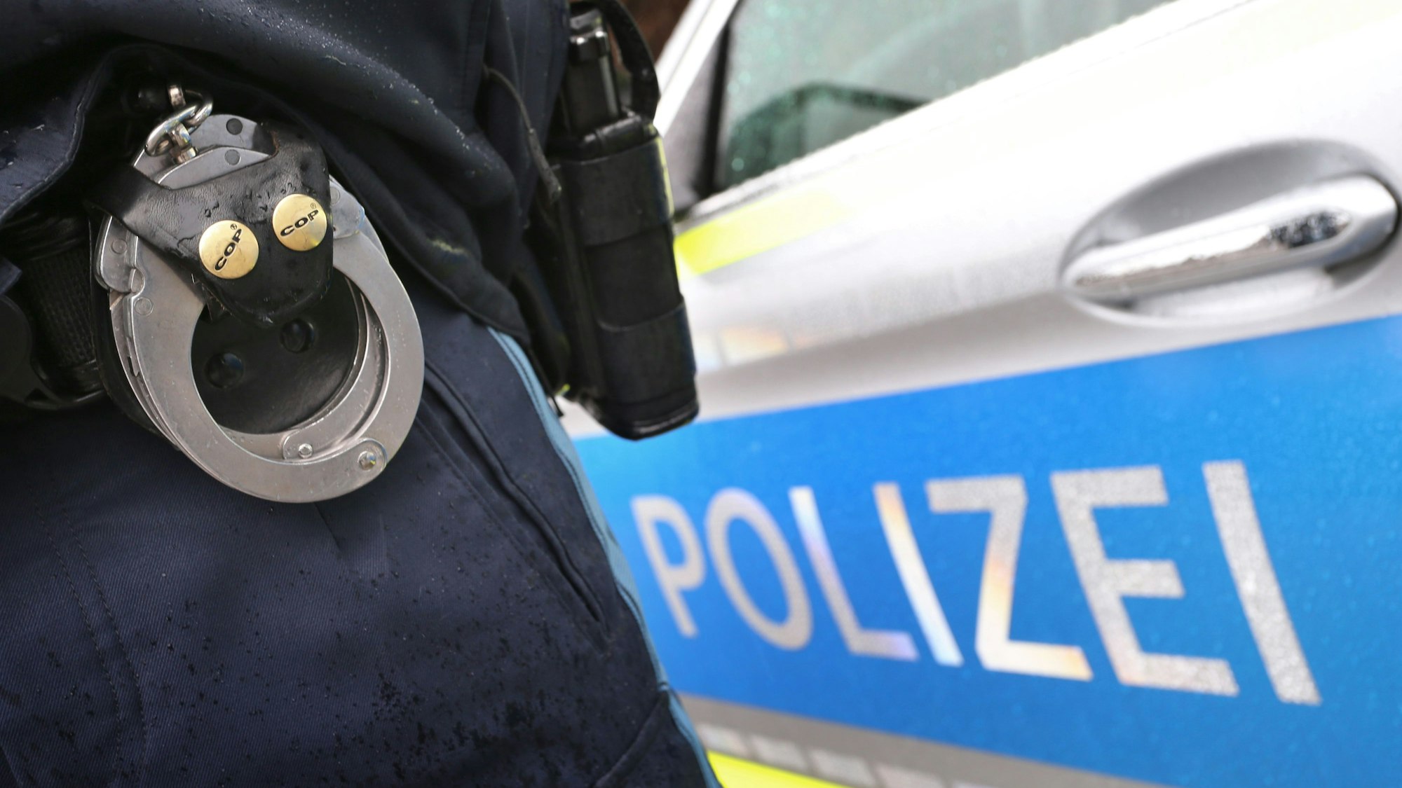Ein Polizist mit Handschellen und einer Pistole am Gürtel steht vor einem Streifenwagen.
