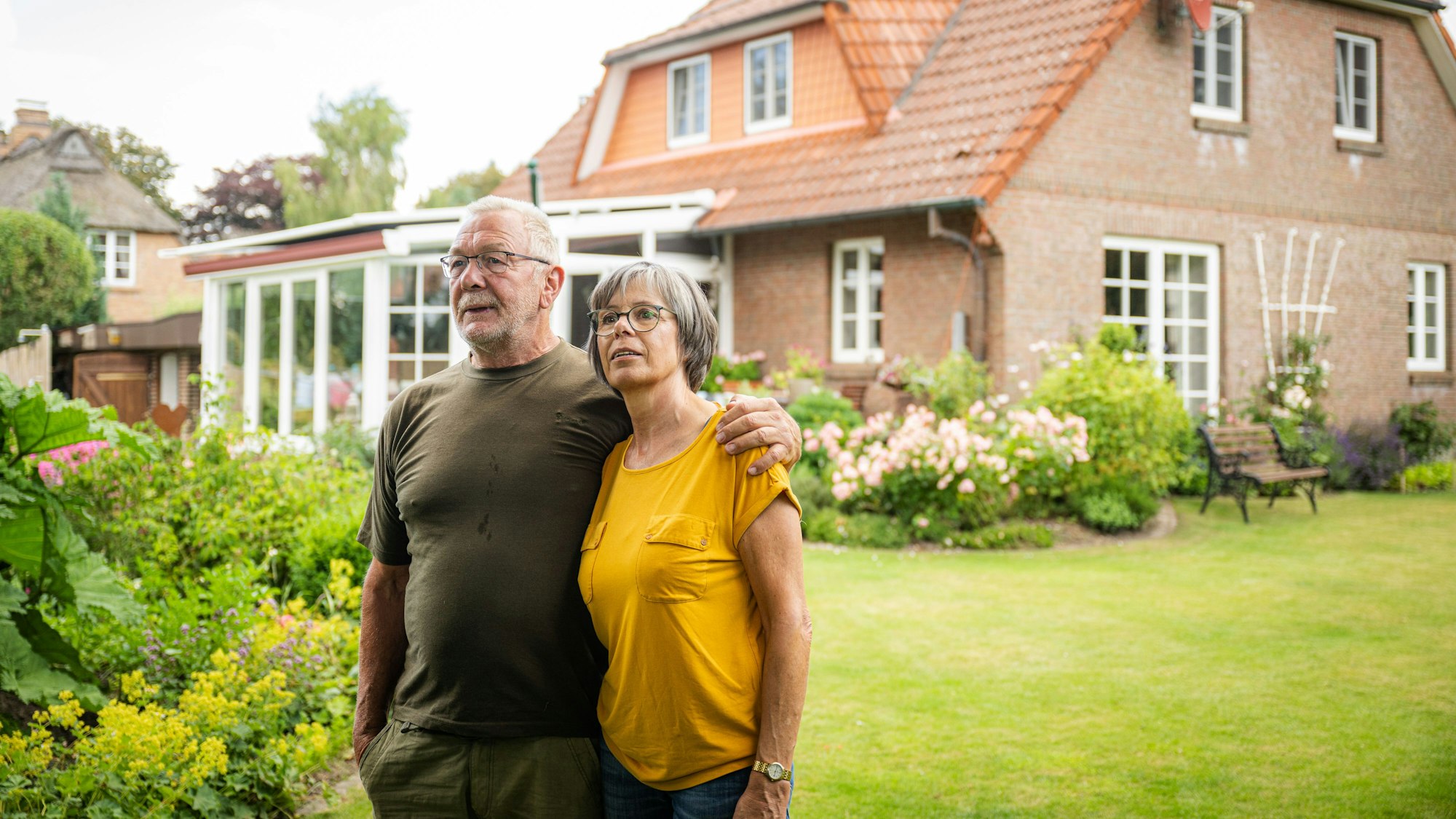 Ein älteres Pärchen steht in einem Garten vor seinem Haus (gestellte Szene).