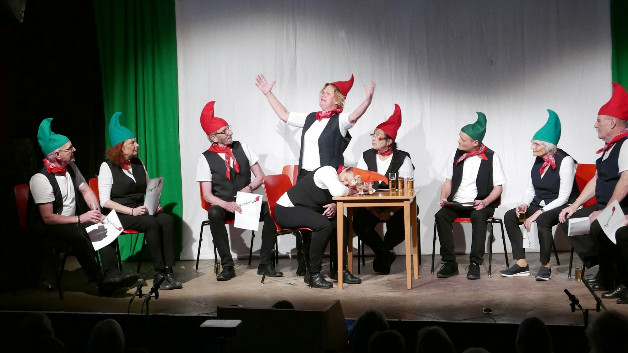 Eine Theatergruppe verkleidet als Heinzelmännchen mit Zipfelmützen sitzt in einem Halbkreis auf der Bühne.