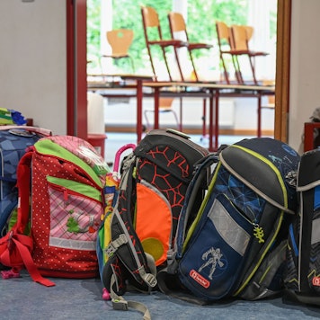Viele bunte Schultornister stehen vor einem Klassenraum, in dem die Stühle hochgestellt sind.