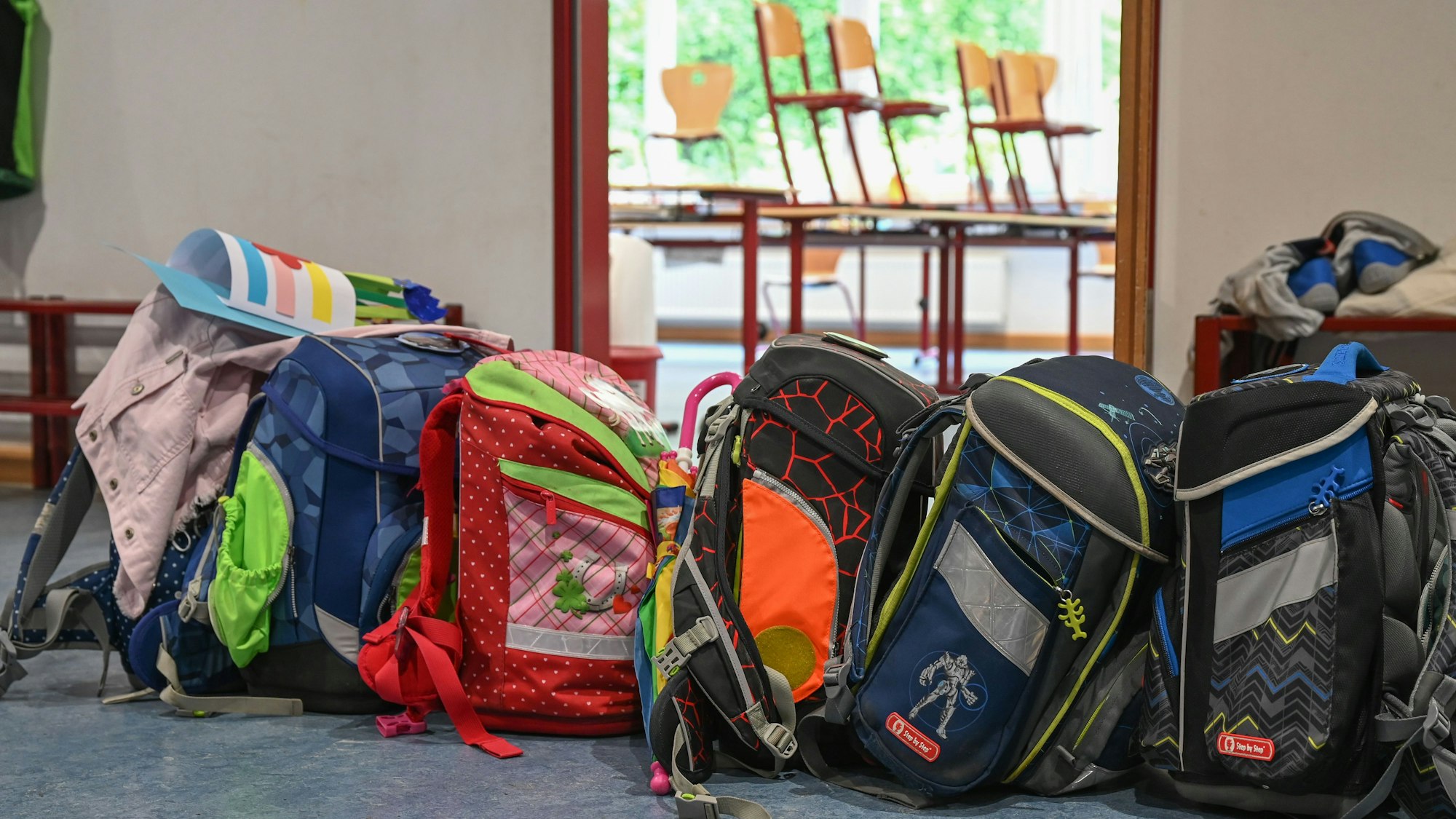 Viele bunte Schultornister stehen vor einem Klassenraum, in dem die Stühle hochgestellt sind.